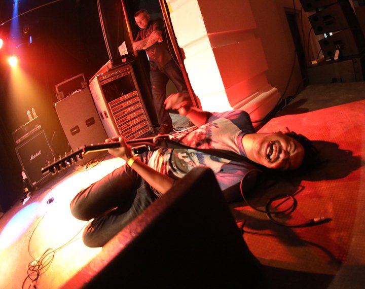 Photo of a man with a guitar, lying on a stage under lights and laughing.