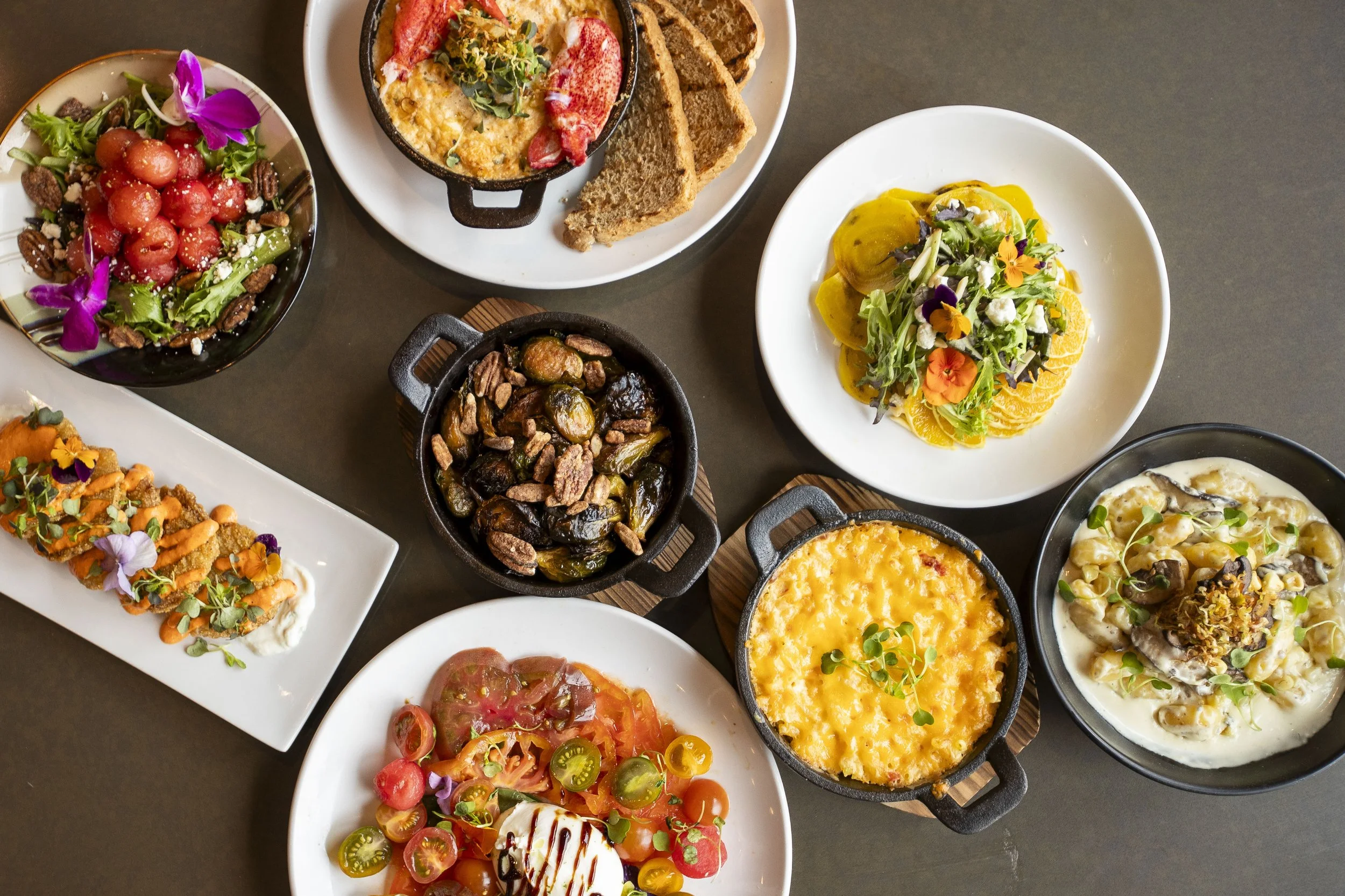Assorted dishes including salads with colorful vegetables and edible flowers, cooked or baked dishes in small cast iron skillets, and a plate of sliced bread with a creamy dip.