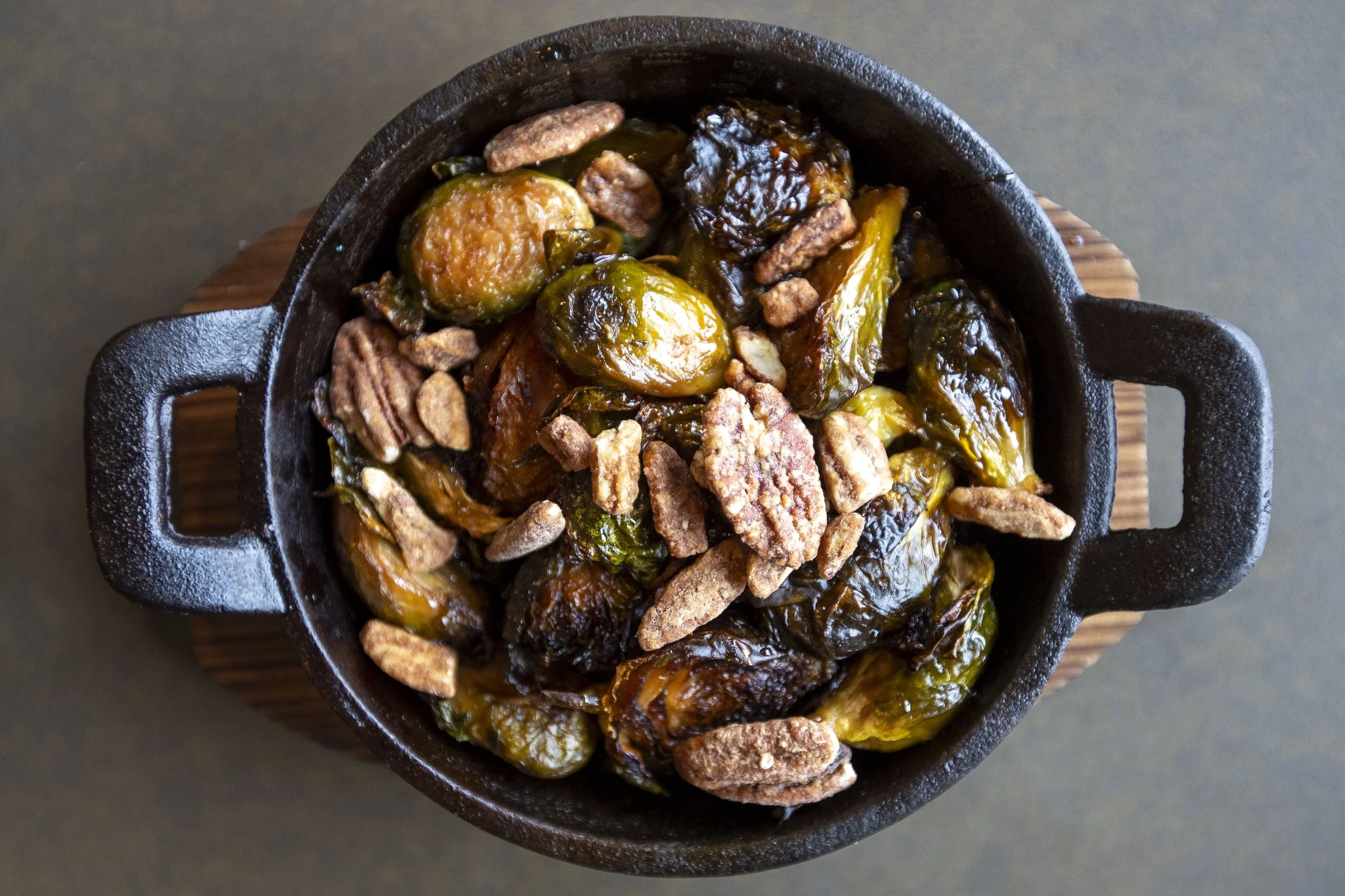 A baked dish in a black cast iron skillet with roasted Brussels sprouts, bacon, and caramelized onions, placed on a wooden trivet.