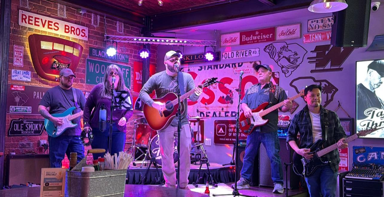 A band of five members performing on a small stage in a bar with colorful signs and brick walls, featuring three guitar players, a bassist, and a singer playing acoustic guitar.