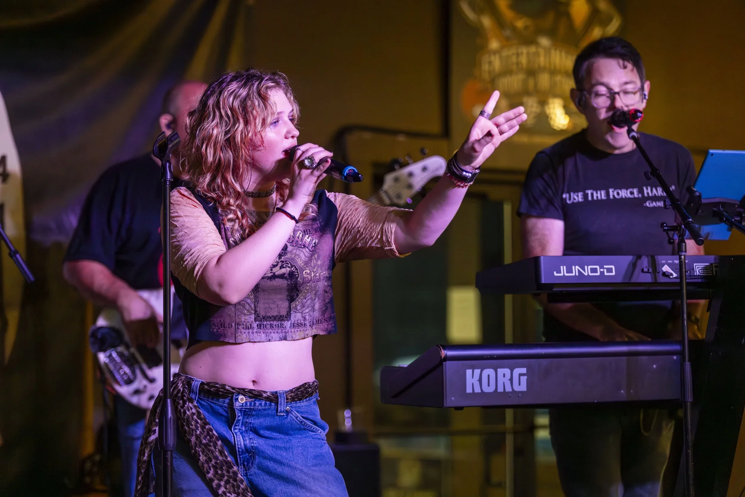 A band of five members performing on a small stage in a bar with colorful signs and brick walls, featuring three guitar players, a bassist, and a singer playing acoustic guitar.