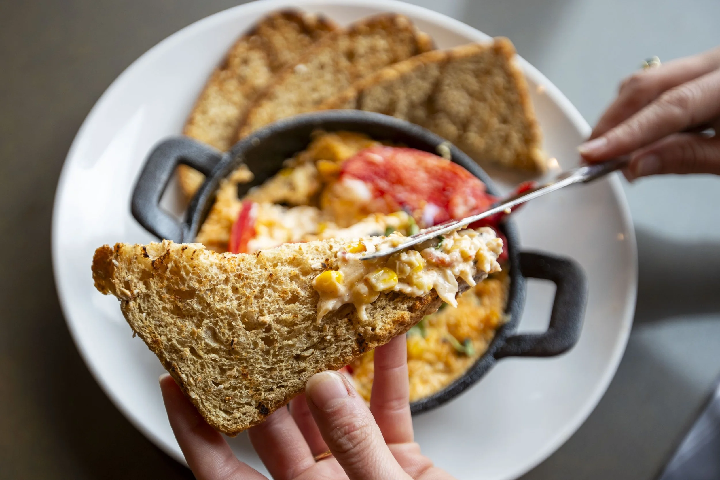 A person holding a slice of bread with a scoop of potato salad, with a plate of nachos topped with cheese, meat, and vegetables in the background.