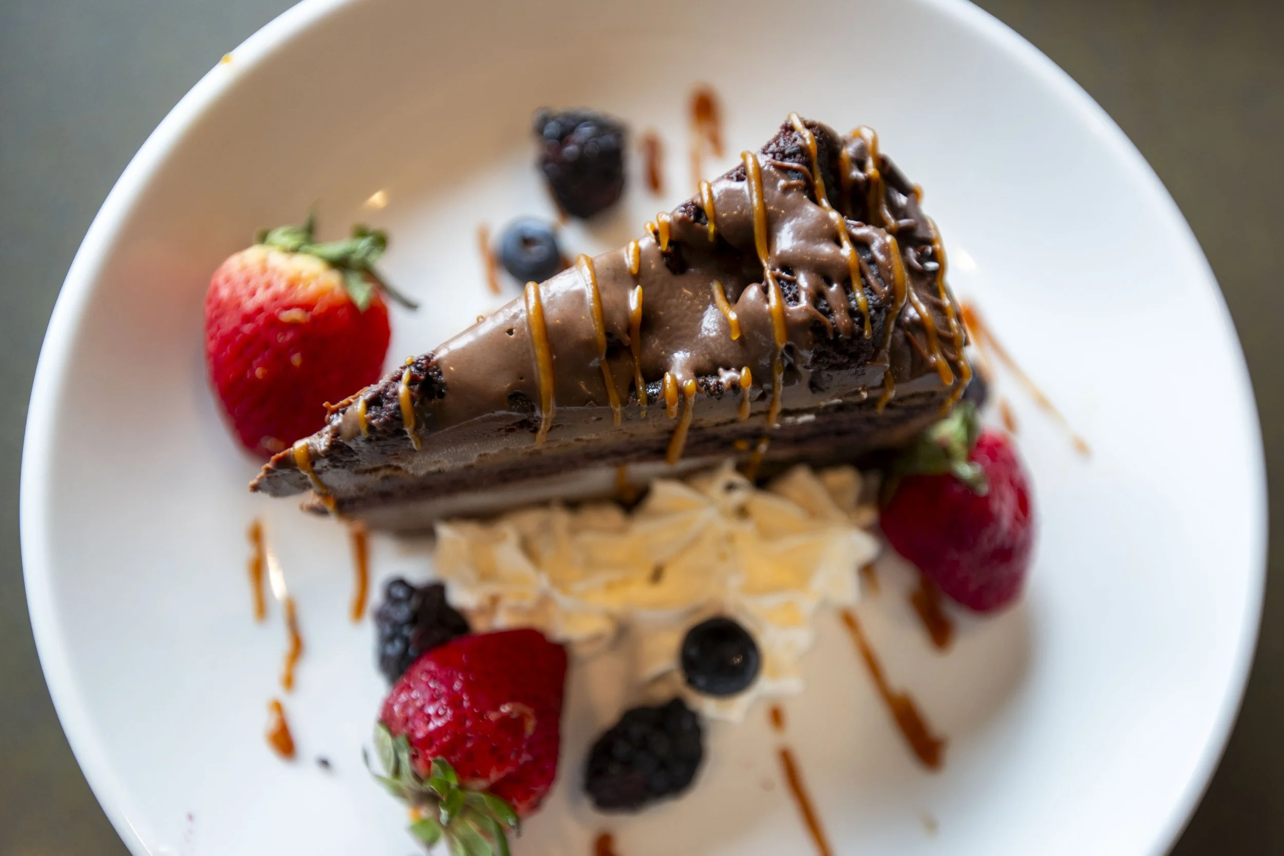 A slice of chocolate cake topped with chocolate drizzle and caramel, garnished with strawberries, blueberries, and blackberries on a white plate.