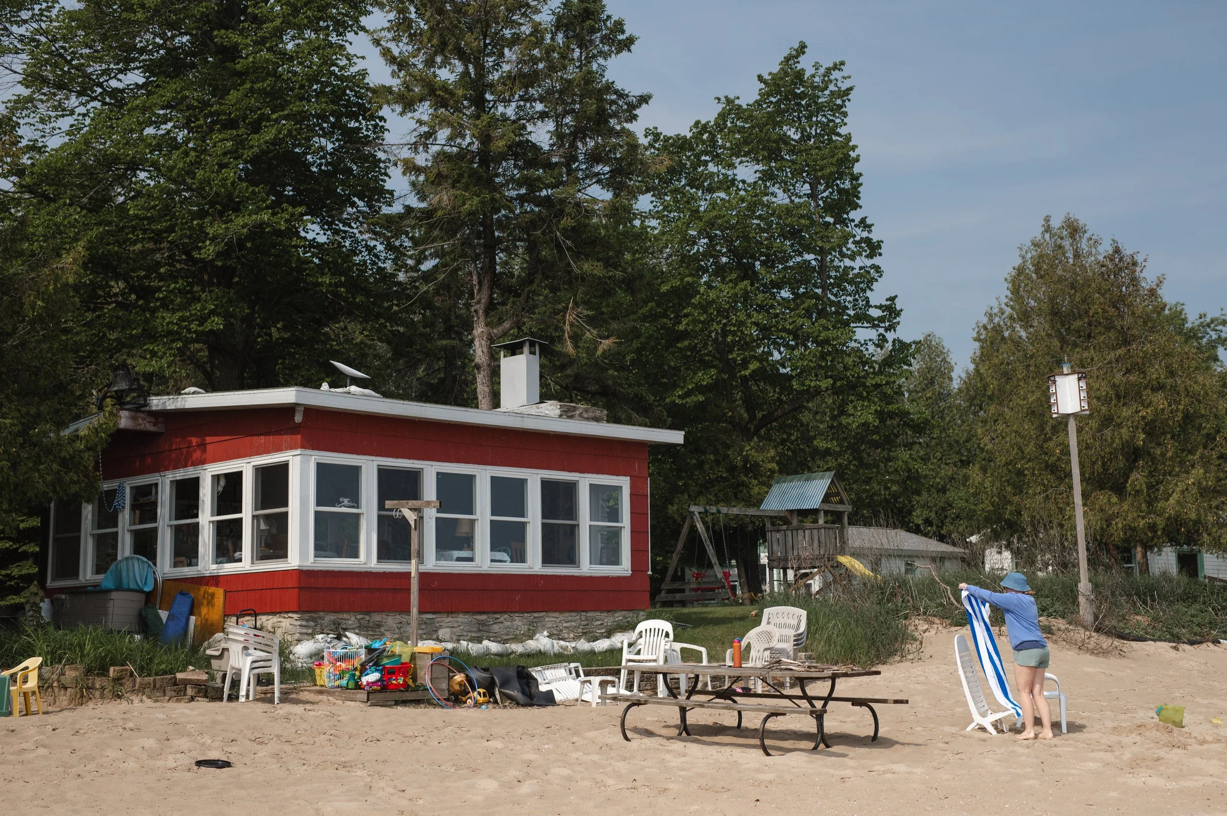 A sandy beach in front of a red cottage with large windows, surrounded by trees. A woman is folding a striped beach chair, with outdoor chairs, a table, and toys scattered nearby.