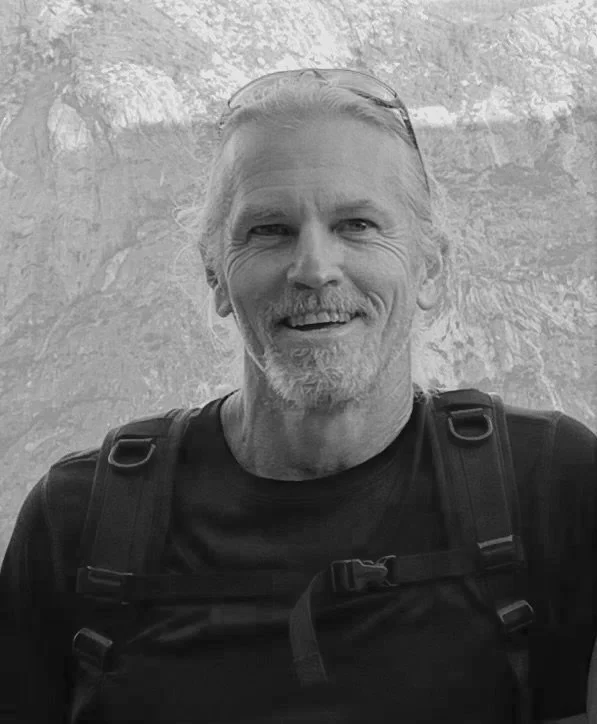A smiling middle-aged man with sunglasses on his head, wearing a black shirt and backpack, outdoors with a mountain background.