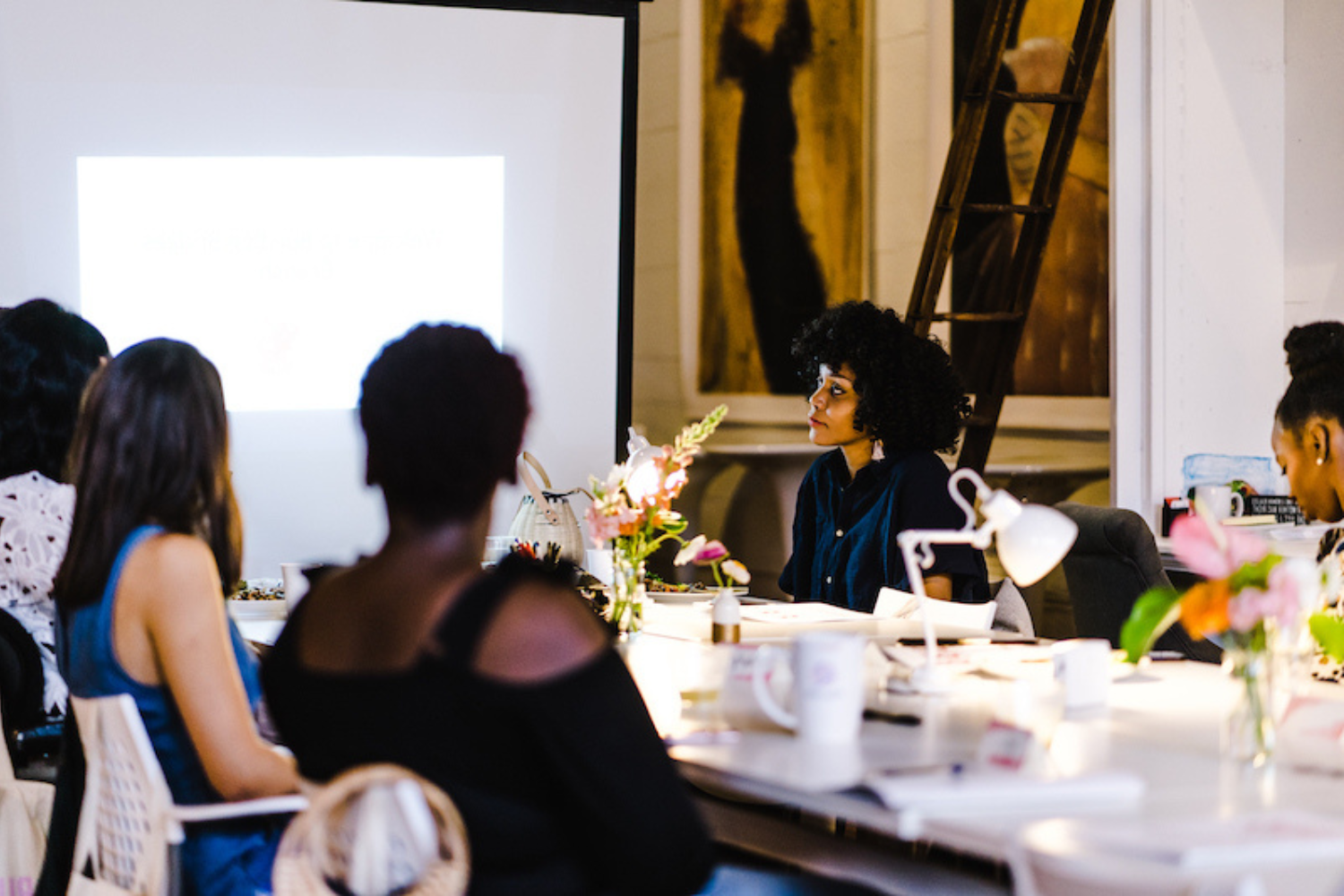 Group of women attending a presentation or workshop in a well-lit room with a woman speaker at the front, projected screen, and floral decorations on the table.