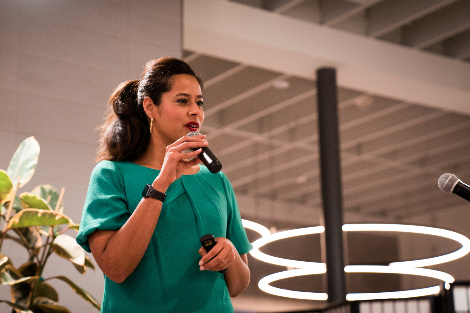 Aparna Rae, a woman in a teal dress speaking into a microphone during an indoor event, with a modern circular light fixture and a plant visible in the background.