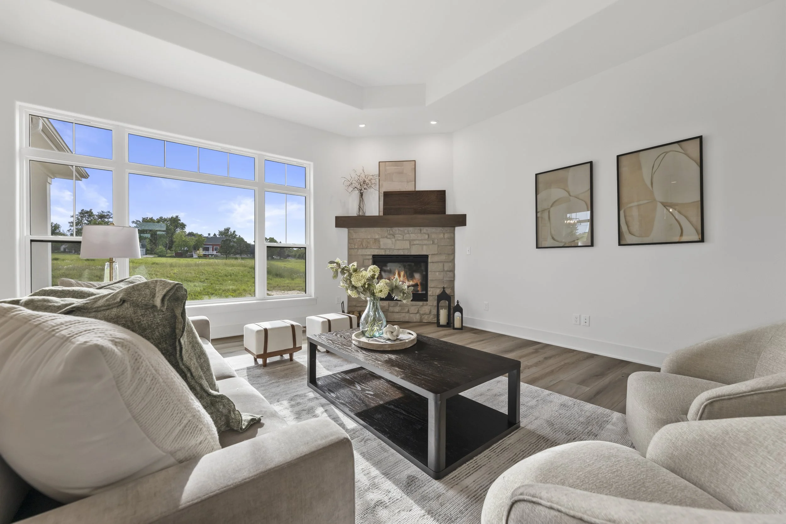 A bright living room with large windows overlooking a green outdoor area. The room has a beige sofa with patterned pillows, a black coffee table with decorative items, a stone fireplace with a wooden mantel, and abstract artwork on the walls.