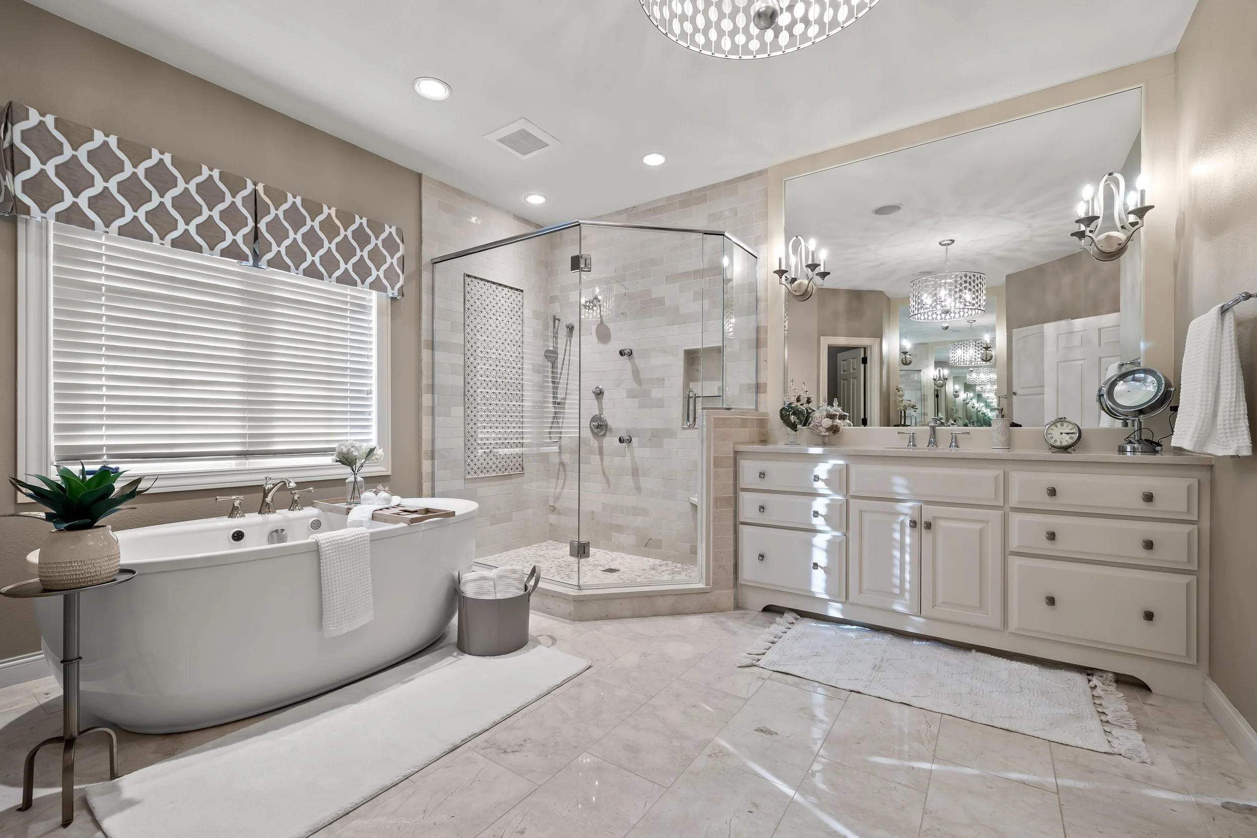 A spacious, elegant bathroom featuring a bathtub with flowers and towels on a small table, a glass shower enclosure with a tiled wall, white cabinetry with silver handles, modern lighting fixtures, a large mirror, and decorative accents.