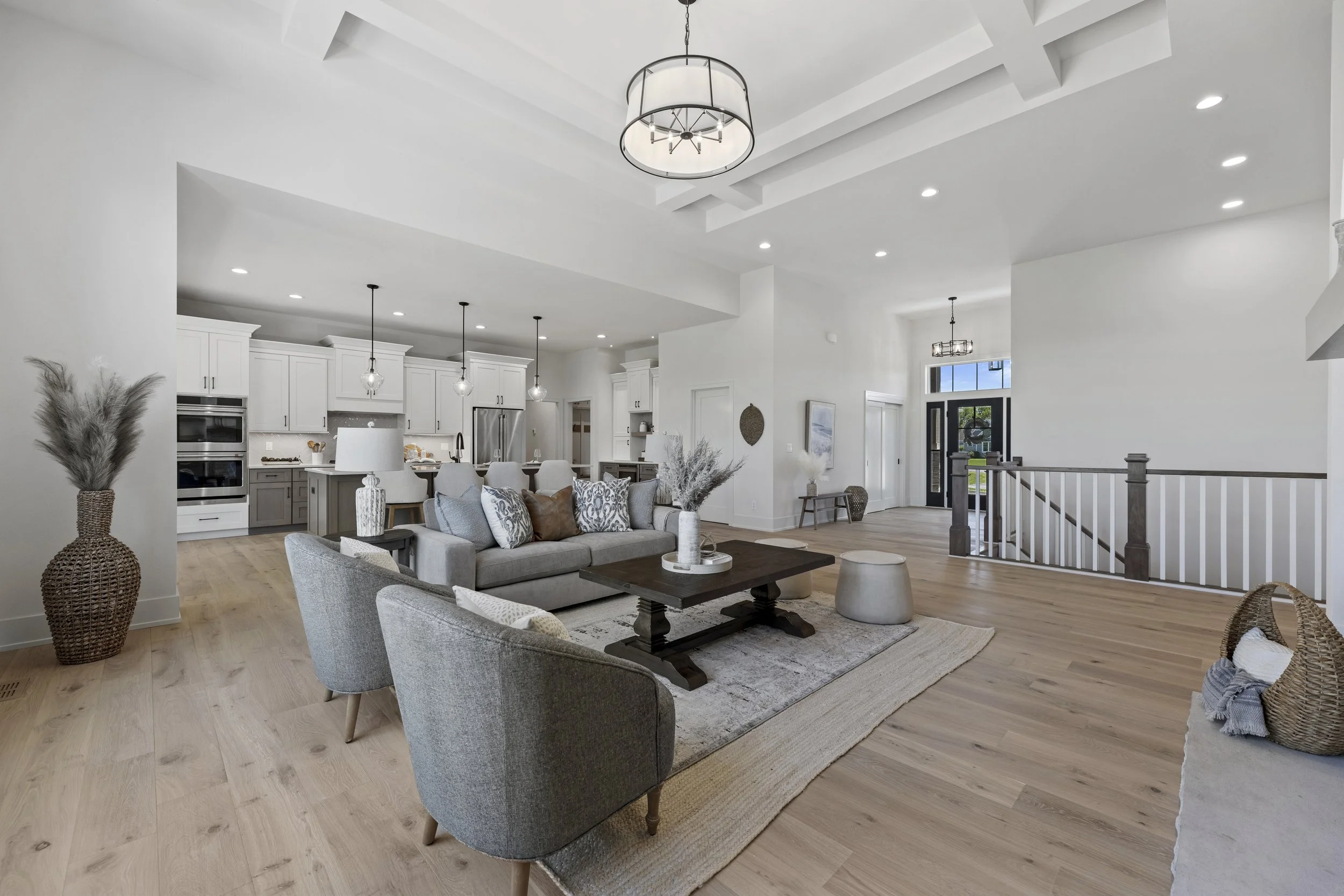 Modern open-concept living room and kitchen with white walls, hardwood flooring, and high ceilings. The living area has a gray upholstered sofa, assorted patterned pillows, and a wooden coffee table. Decor includes woven vases with dried plants and a