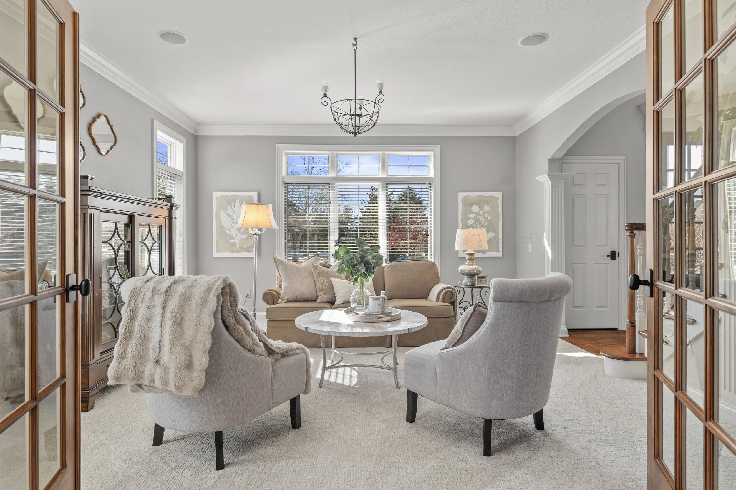Living room with beige and gray furniture, a coffee table with a plant and decorative items, large windows with white blinds, and framed artwork on the walls.