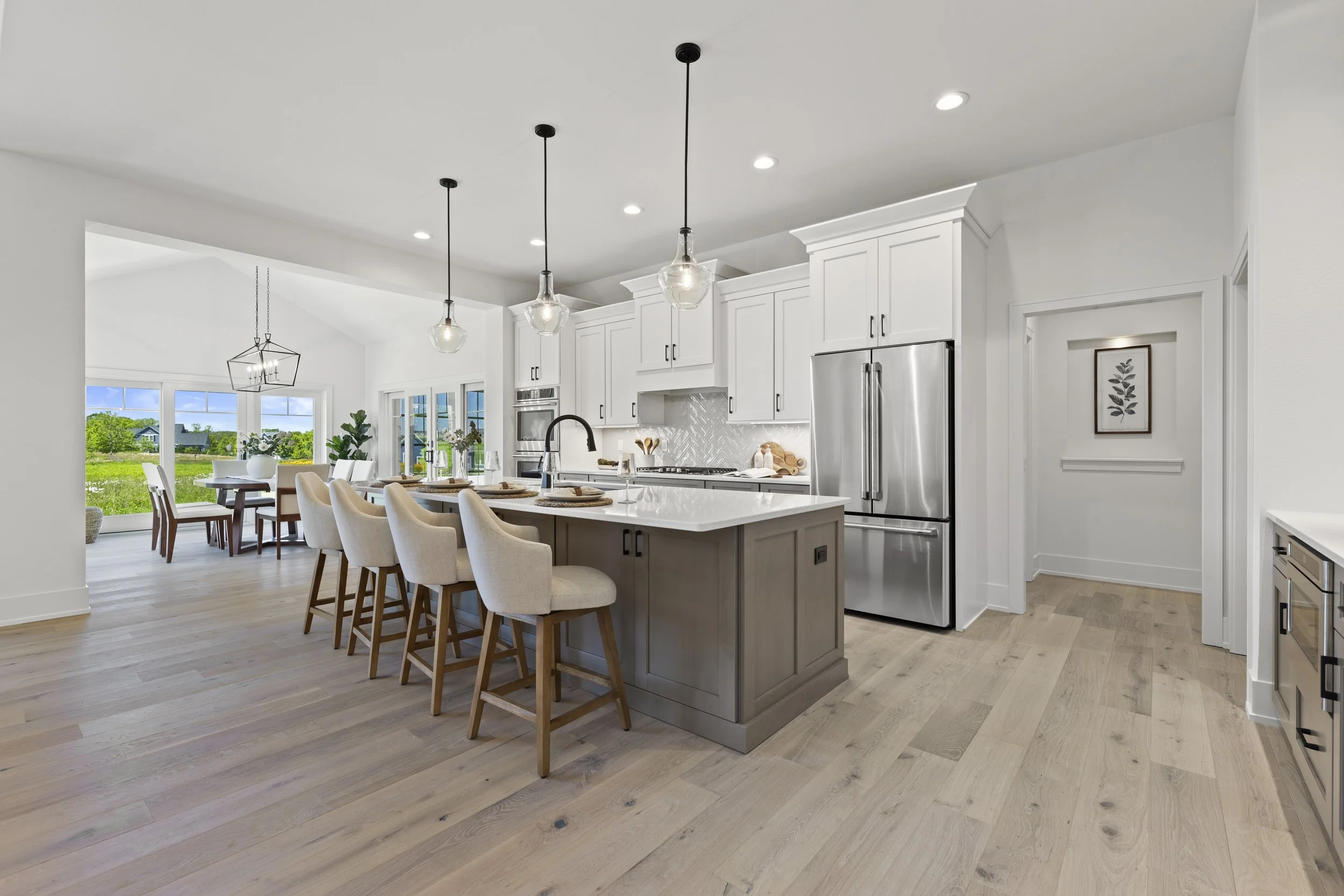 Modern open-concept kitchen with white cabinets, stainless steel appliances, island with beige chairs, pendant lights, dining area with white chairs, large windows with a view of greenery outside.