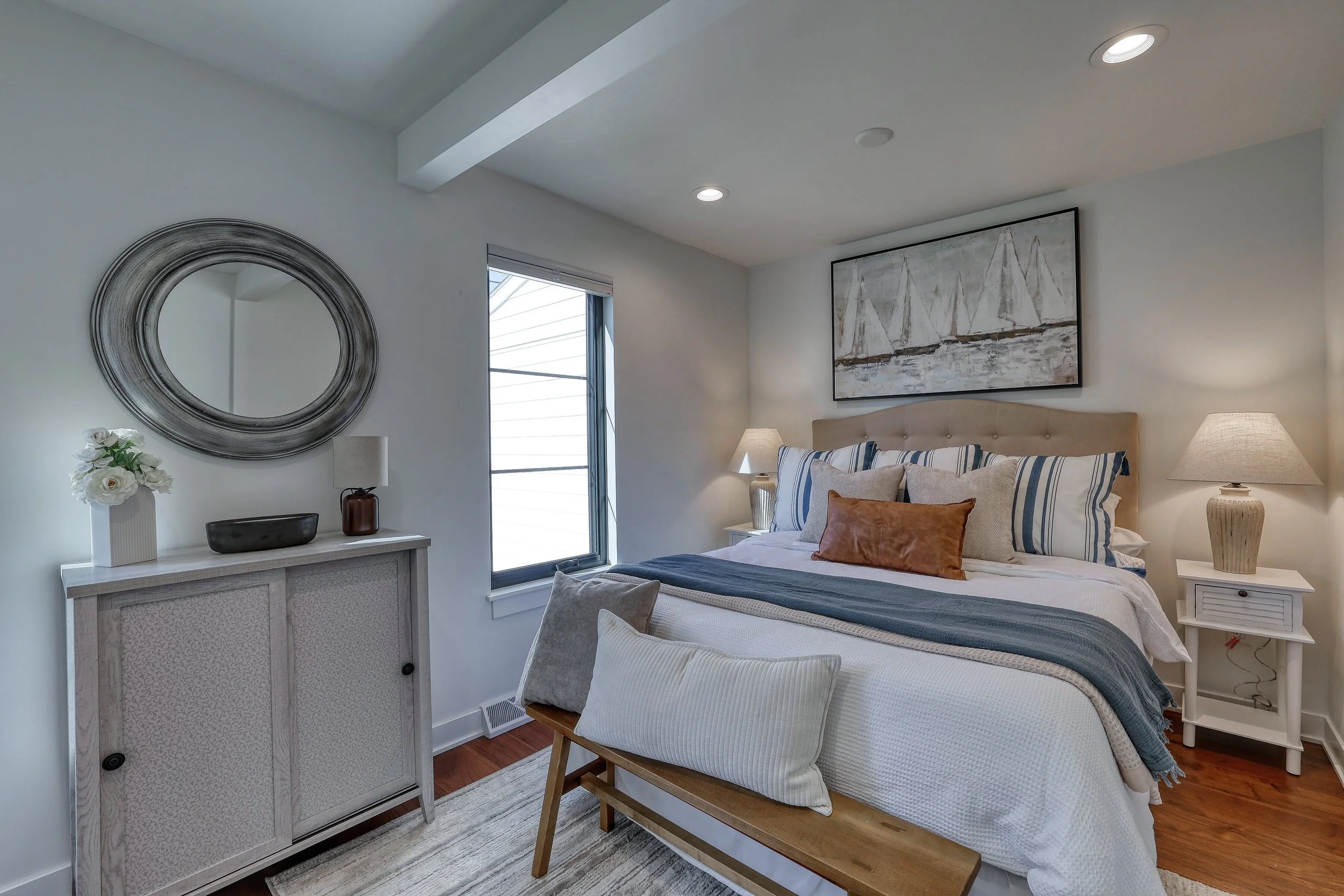 A cozy bedroom with a large bed, striped pillows, and a beige headboard. There are two nightstands with lamps, and a large abstract painting of sailboats hangs above the bed. A decorative mirror and flowers are on a white cabinet near a window with b