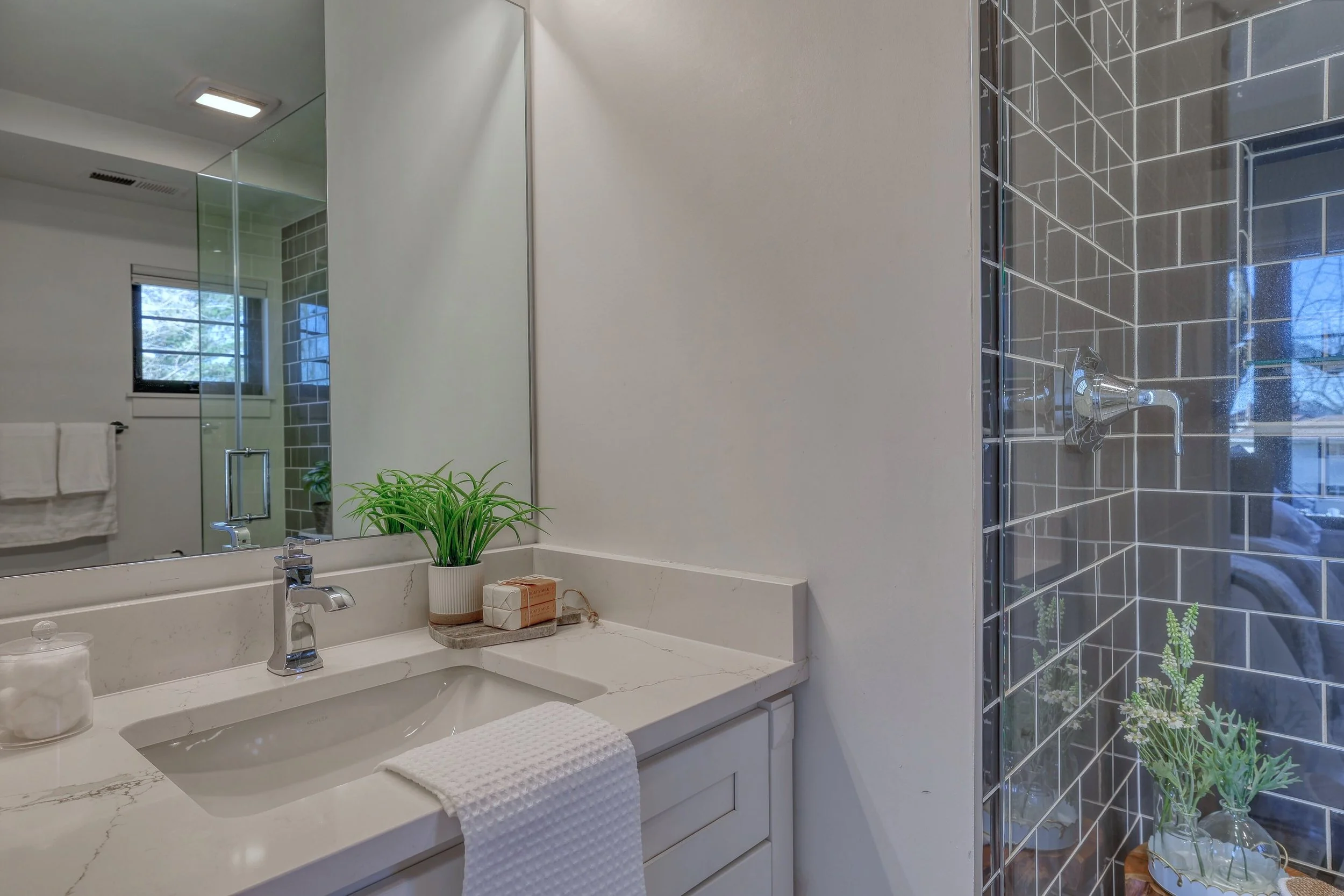 Modern bathroom with white countertop, sink, and a large mirror. Small potted plant and toiletries on the counter. Part of a shower with glass door and gray tiles with white grout. Window showing trees outside.