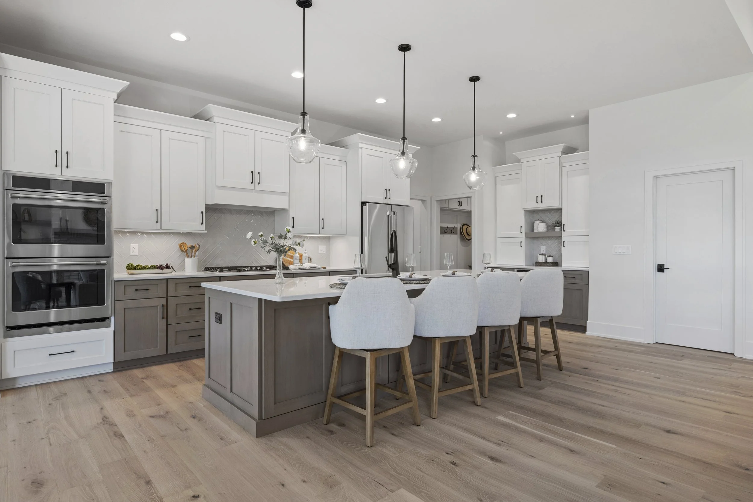 Modern kitchen with white cabinets, a kitchen island with a gray base, bar stools with white cushions, pendant lights, and stainless steel appliances, including a double oven and a refrigerator. The kitchen has light wood flooring and a minimalist de