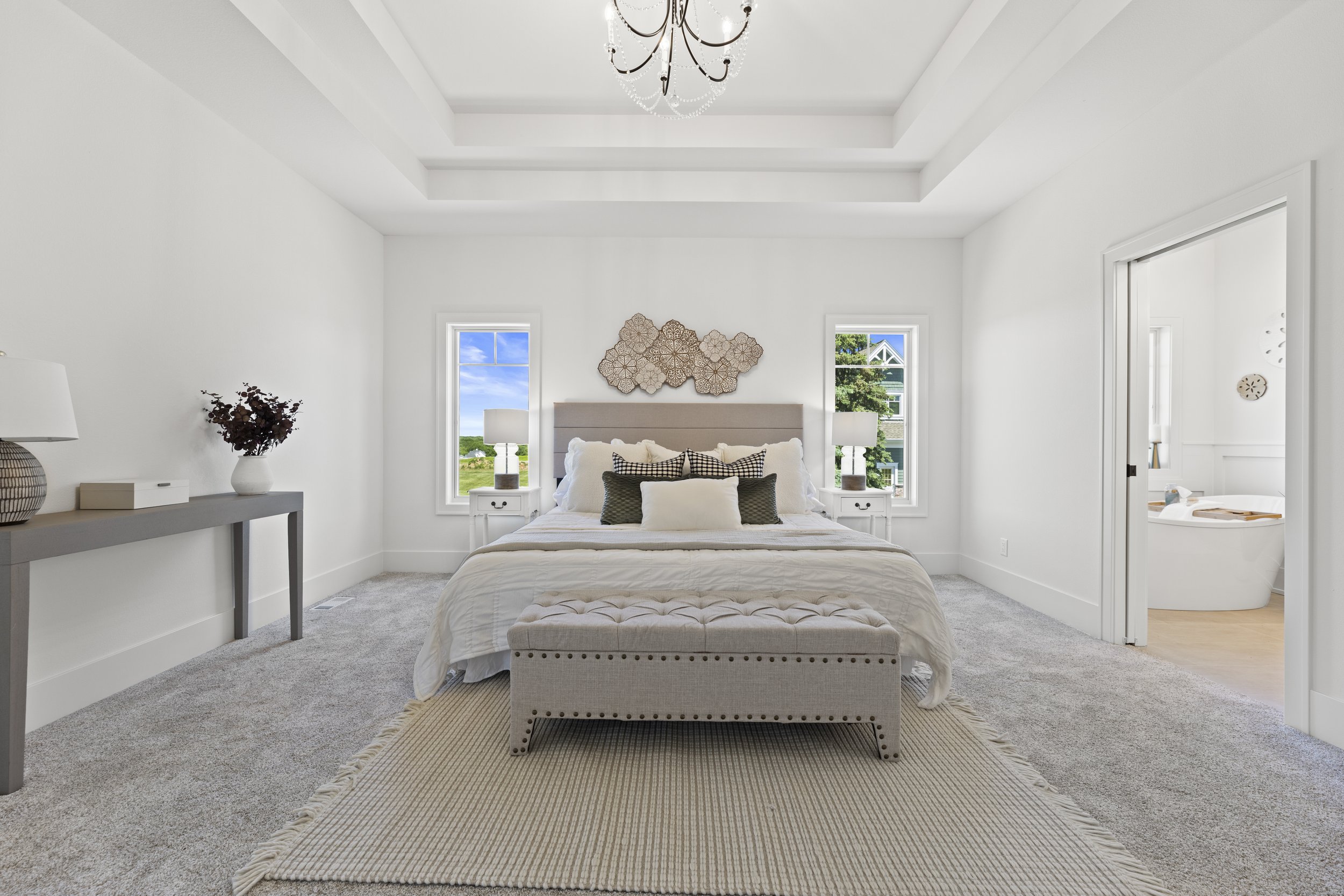 A bright modern bedroom with white walls and ceiling, a chandelier, and two windows showing a view of green trees and blue sky. The bed has white bedding with multiple pillows, flanked by two small white nightstands with lamps. A beige upholstered be