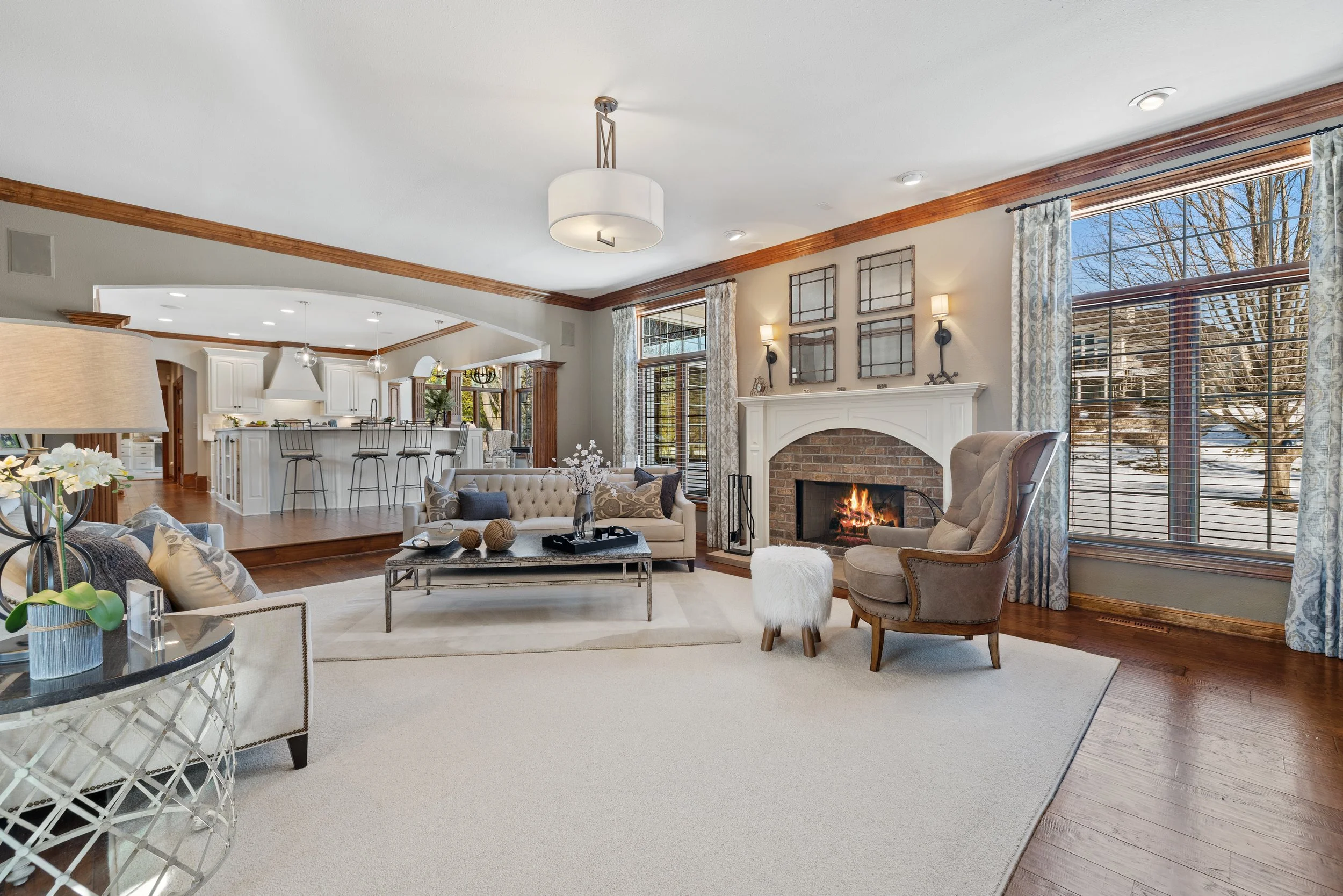 Living room with fireplace, large windows with curtains, sofa with cushions, armchair, coffee table, and decorative items, open to kitchen area.