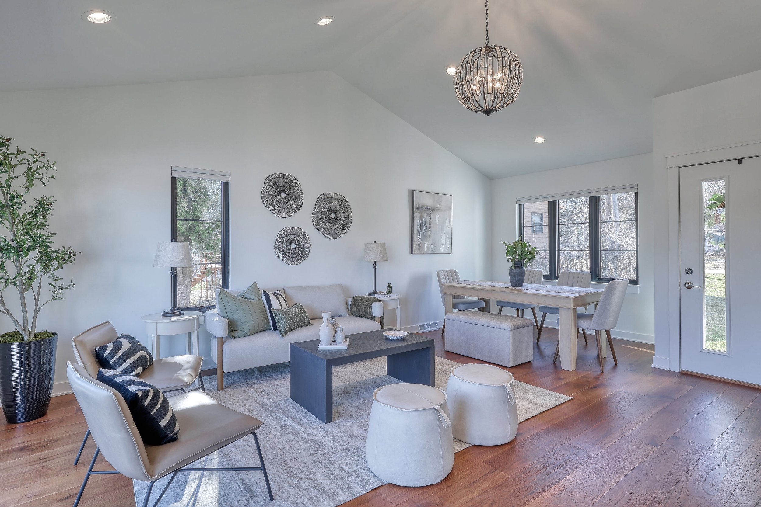 Living room with white walls, large windows, and a ceiling light fixture. It features a beige sofa with multiple throw pillows, two white side tables with lamps, a dark rectangular coffee table, and a dining area with a wood table and beige chairs. T