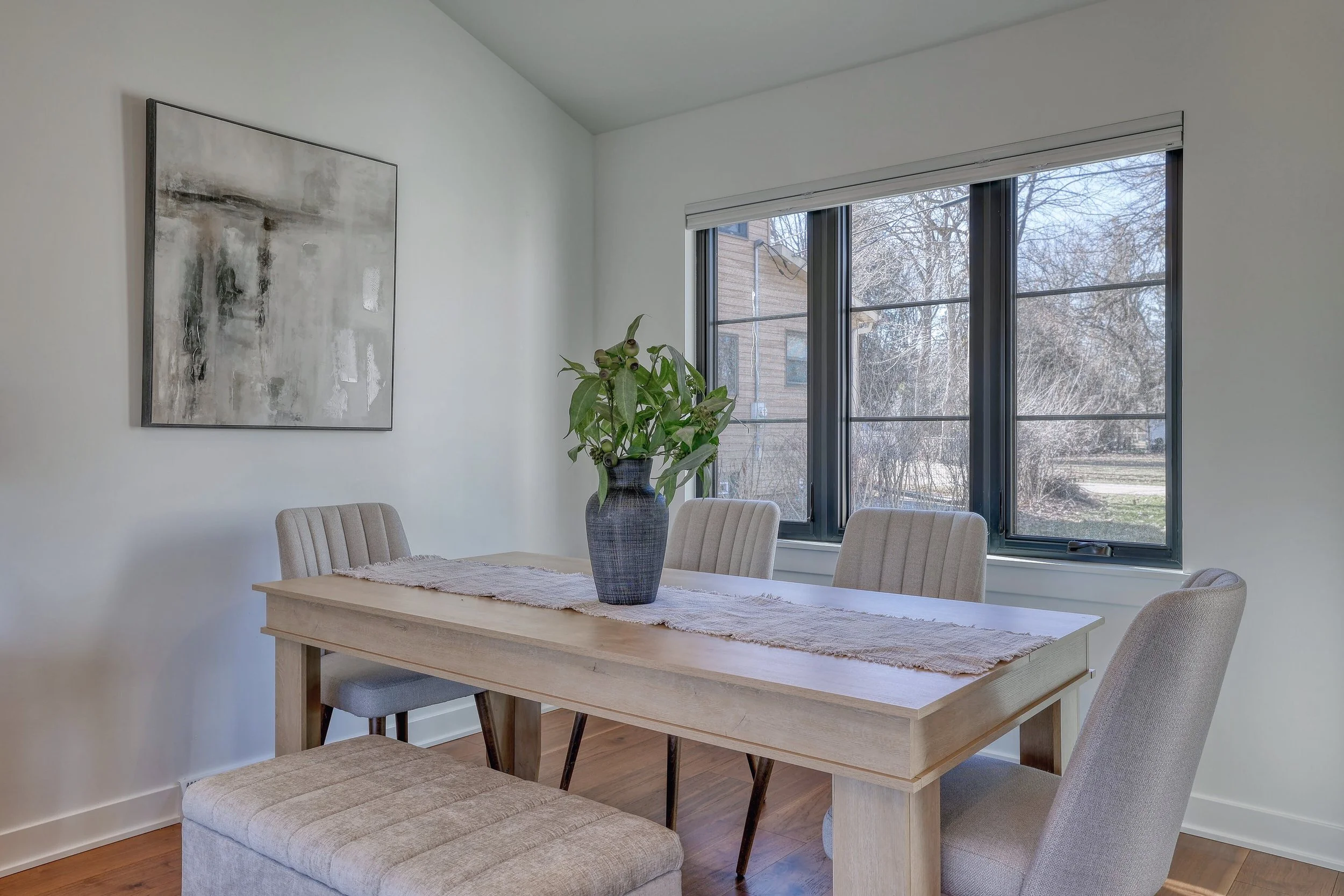A dining room features a light-colored wooden table with a runner and a large potted plant in a ceiling-high window. There is a large abstract painting on the white wall and a beige upholstered bench at the end of the table, with four matching chairs
