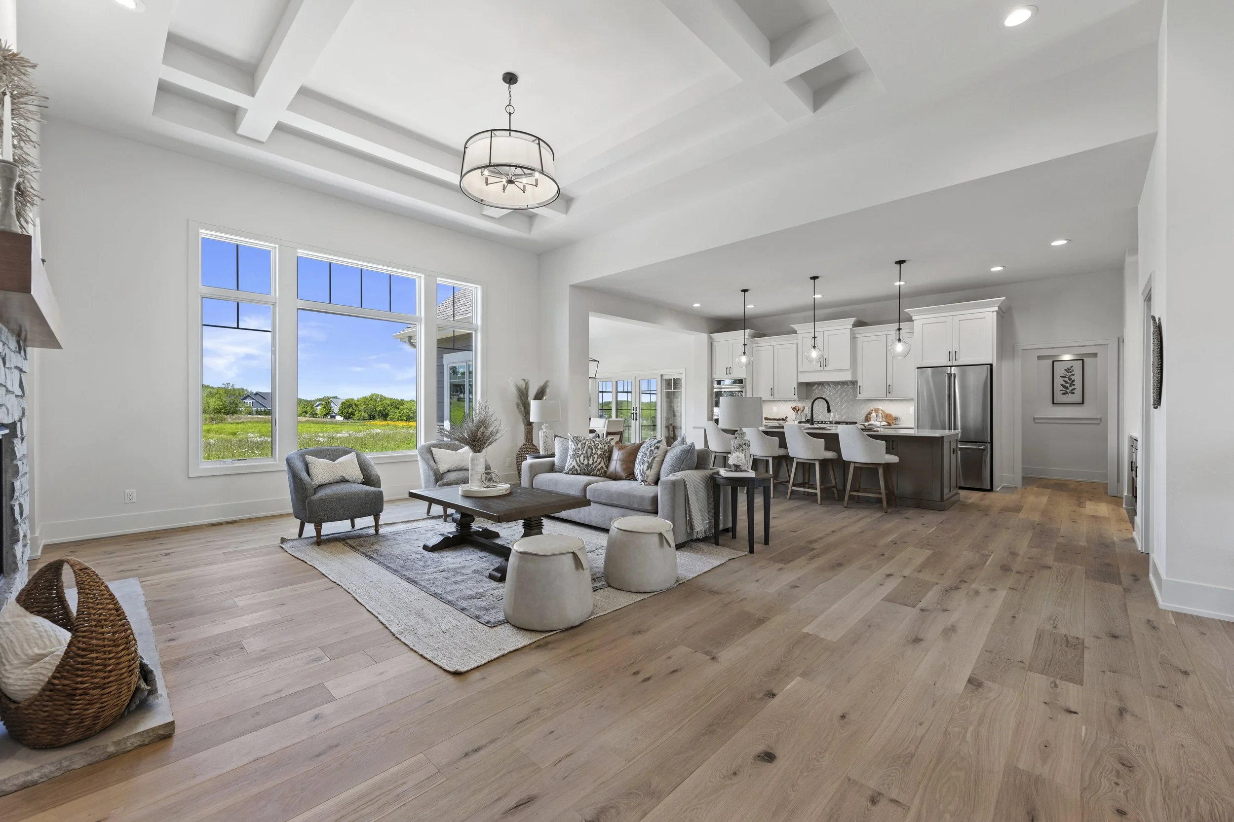 Bright open living room with large windows showcasing green landscape, featuring a cozy seating area with a sofa, armchair, and coffee table, connecting to a modern kitchen with white cabinetry and pendant lighting.