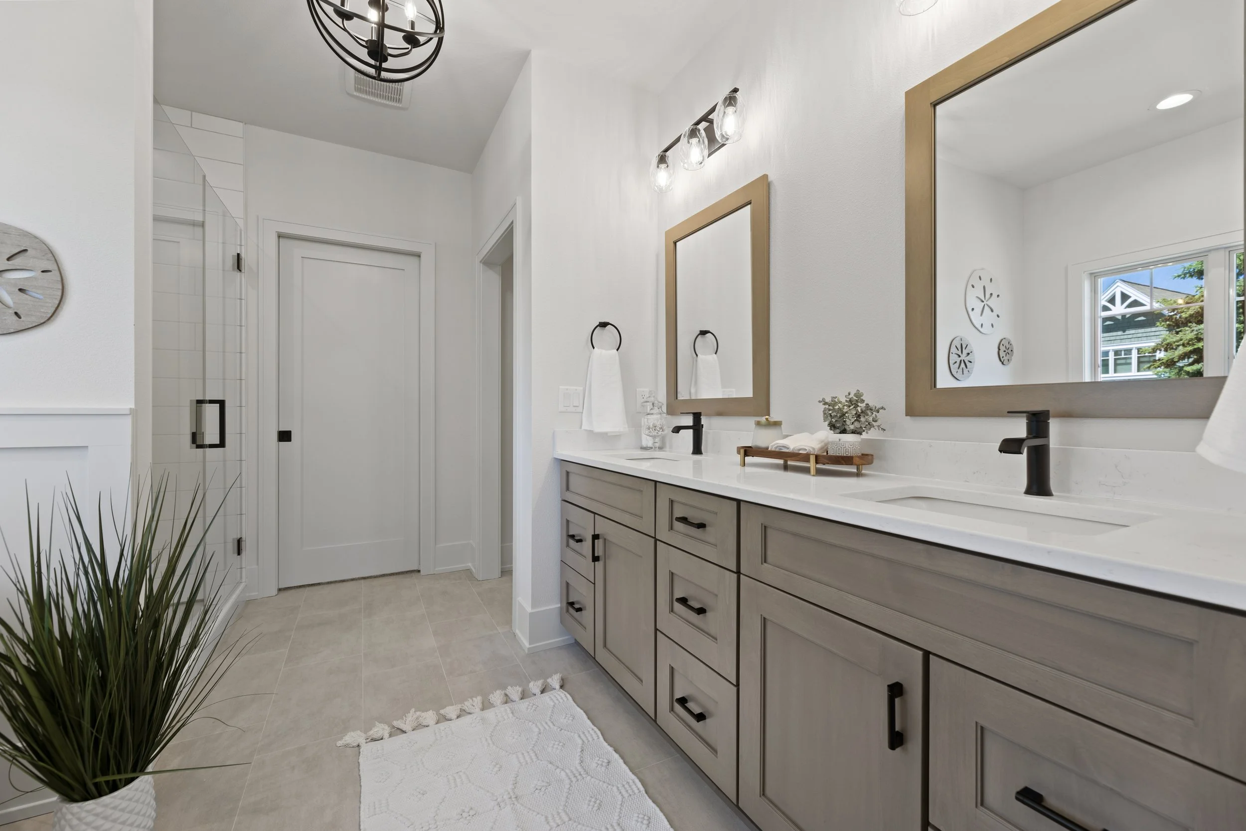 A modern bathroom with a double vanity featuring a white countertop and light gray cabinets with black hardware. Two large mirrors with wooden frames are positioned above the sinks, and a three-light vanity fixture with glass shades is mounted above 