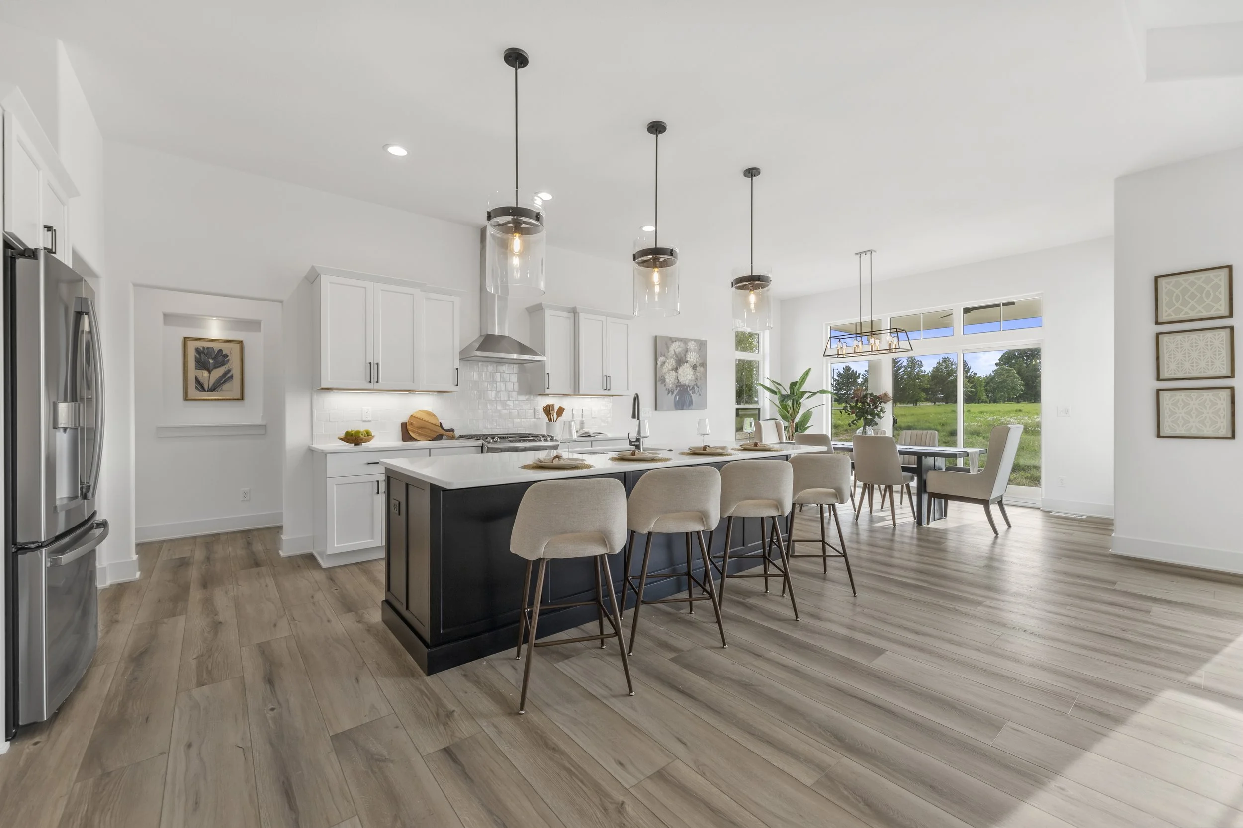 Modern white kitchen with island, beige barstools, dining table, large window overlooking green fields, pendant lights, and wooden flooring.