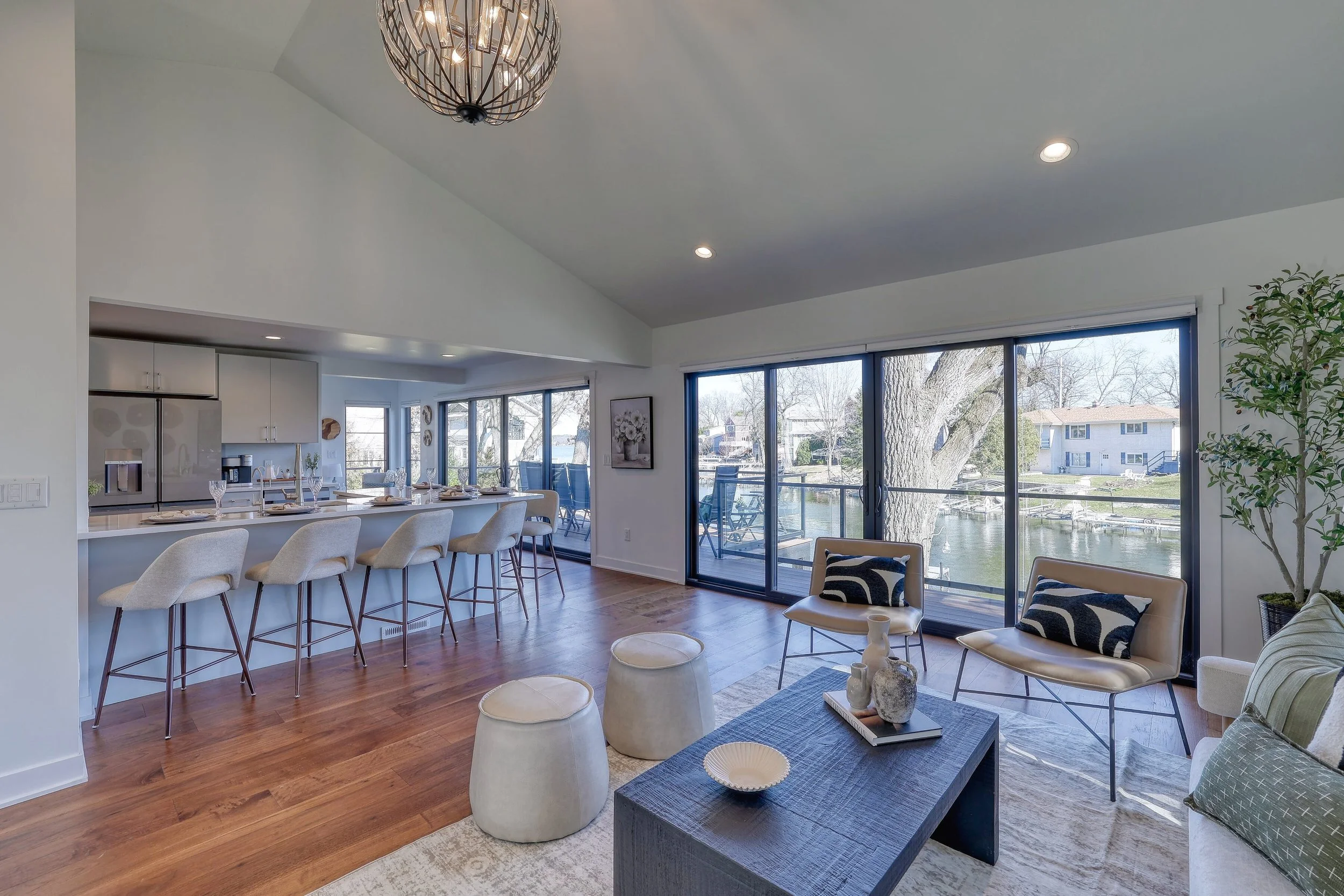 Living room and kitchen with large windows overlooking a river and trees outside. Modern furniture, hardwood floors, and a decorative chandelier.