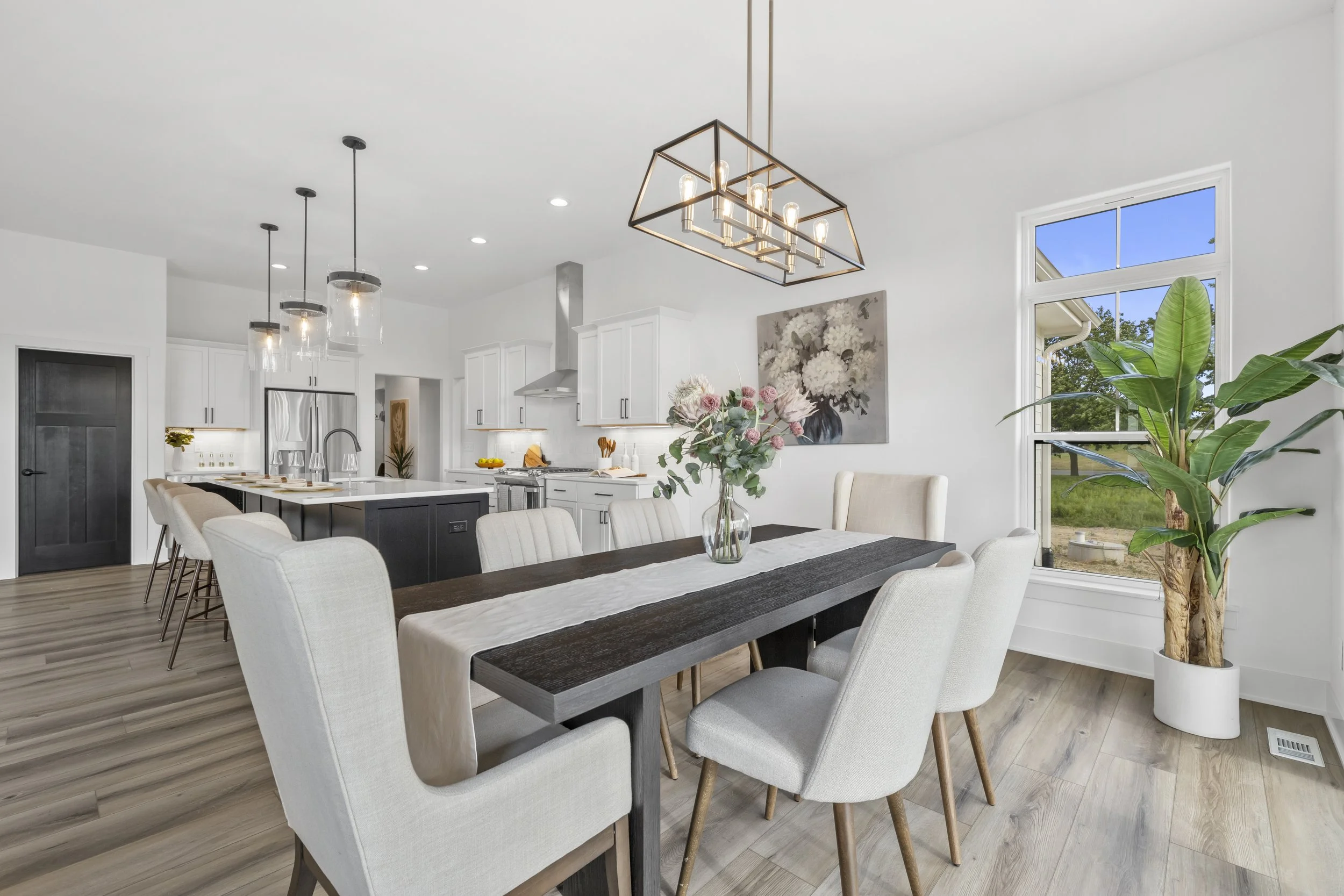 Bright, modern dining and kitchen area with white cabinets, stainless steel appliances, a dark dining table with light-colored chairs, large windows with a view of trees, and indoor plants.