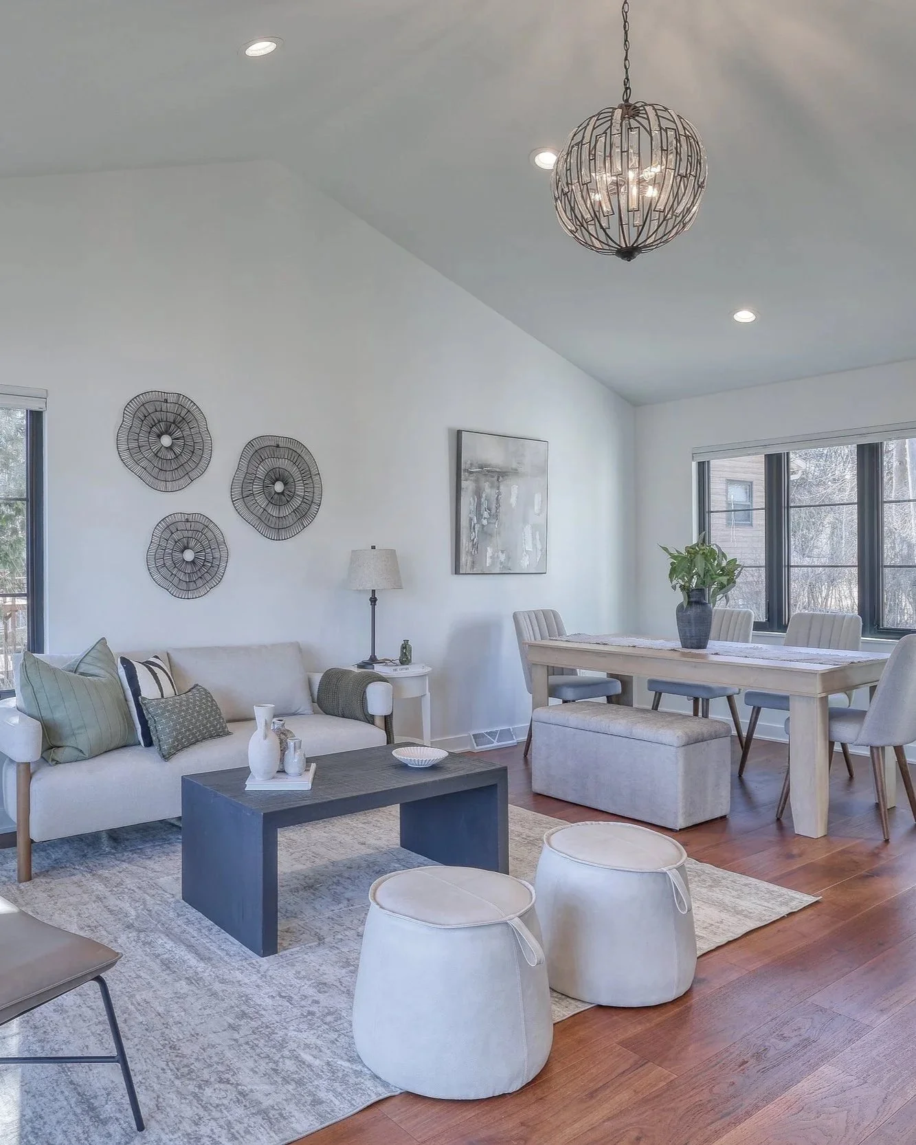 Bright living room with white walls, a window with blinds, a beige sofa decorated with pillows, a black coffee table with vases, a side table with a lamp, wall art, a dining table with chairs and a potted plant, and a chandelier.