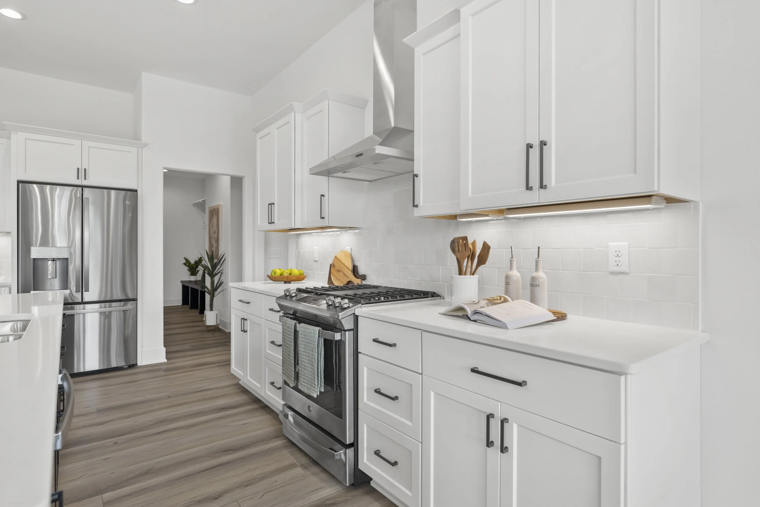 Modern white kitchen with stainless steel refrigerator, gas stove, white cabinets with black handles, and a white countertop with a book, olive oil bottles, and a bowl of lemons. There is wood flooring and a small hallway with a plant and artwork in 