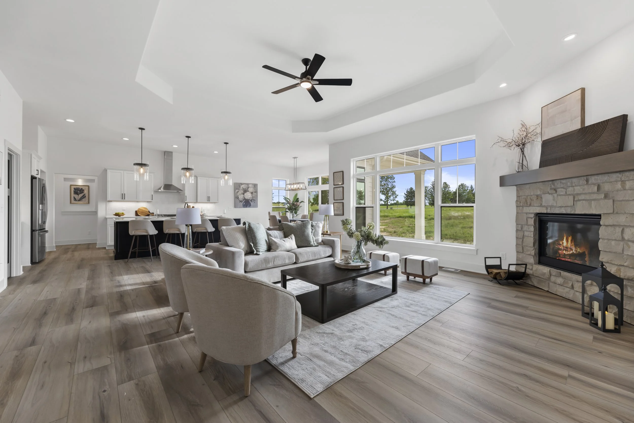 Bright living room with large windows showing a grassy outdoor scene, white walls, a stone fireplace with a fire, modern ceiling fan, and cozy furniture.