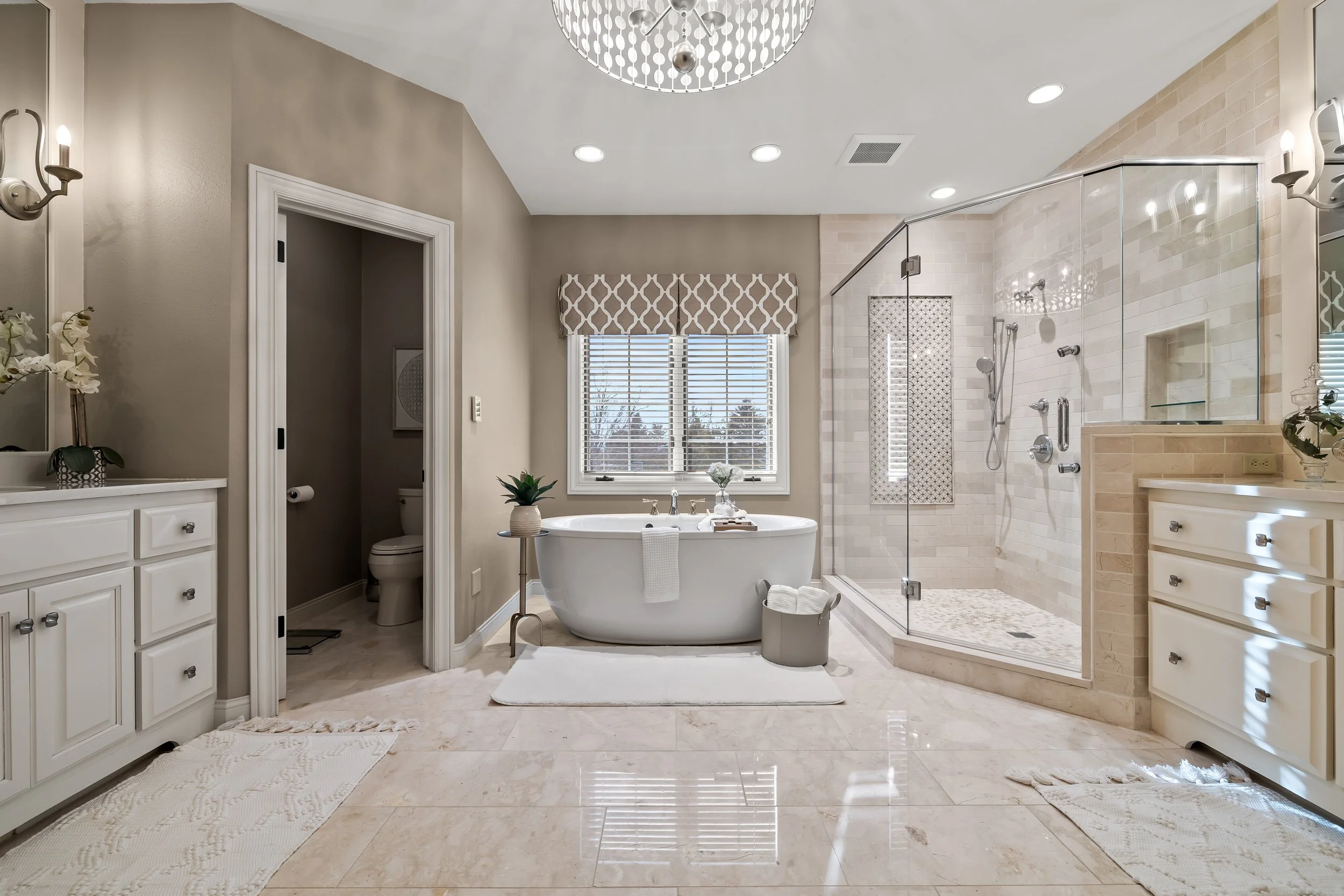 A spacious, well-lit bathroom featuring a bathtub, a glass-enclosed shower, white cabinetry, and a window with a beige and white valance. The room has beige tile flooring and neutral-colored walls.