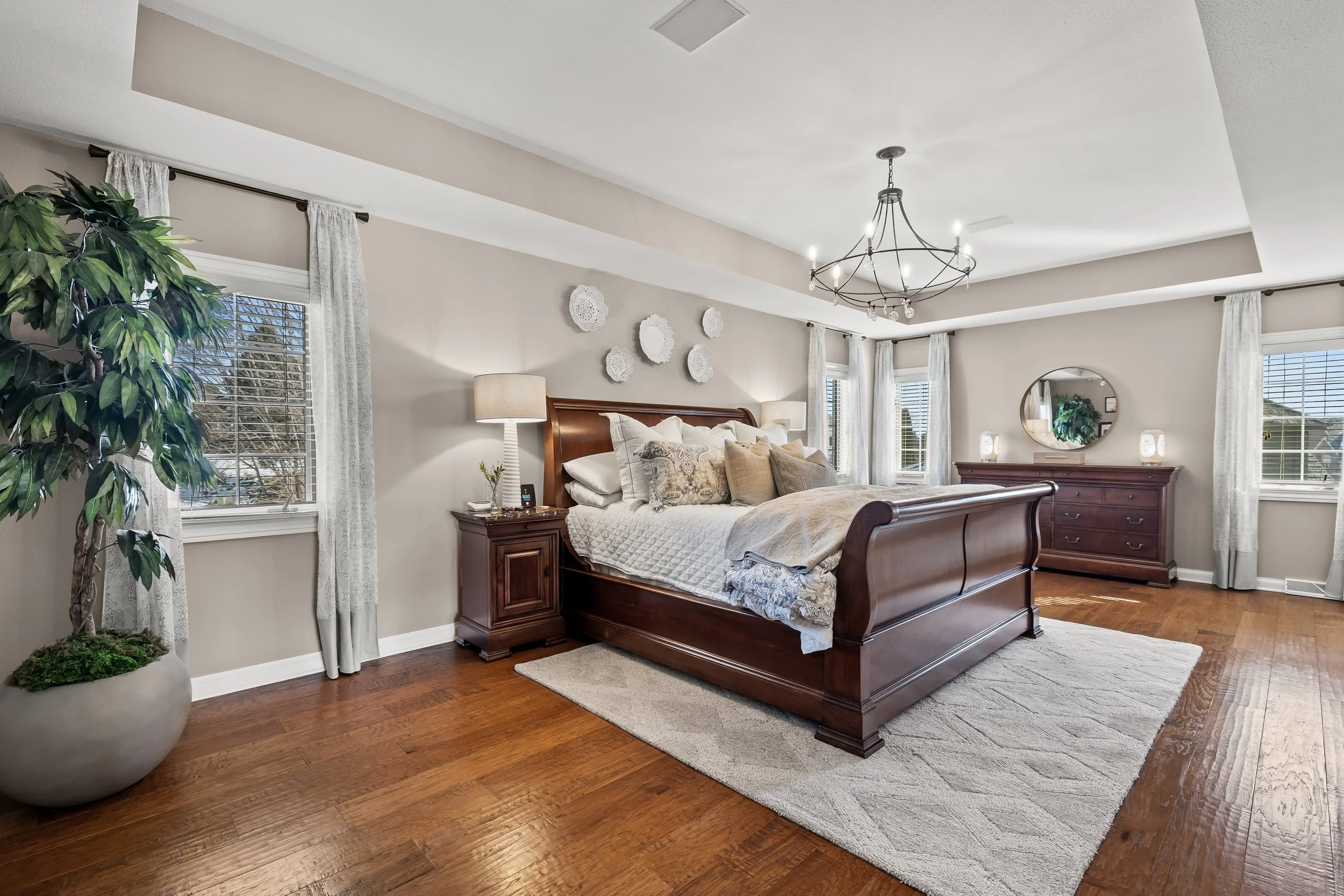 A bedroom with a wooden bed, white bedding, beige and patterned throw pillows, wooden nightstands with lamps, a dresser with a mirror, and several windows with white curtains. There is a large green potted plant in the corner and white decorative wal