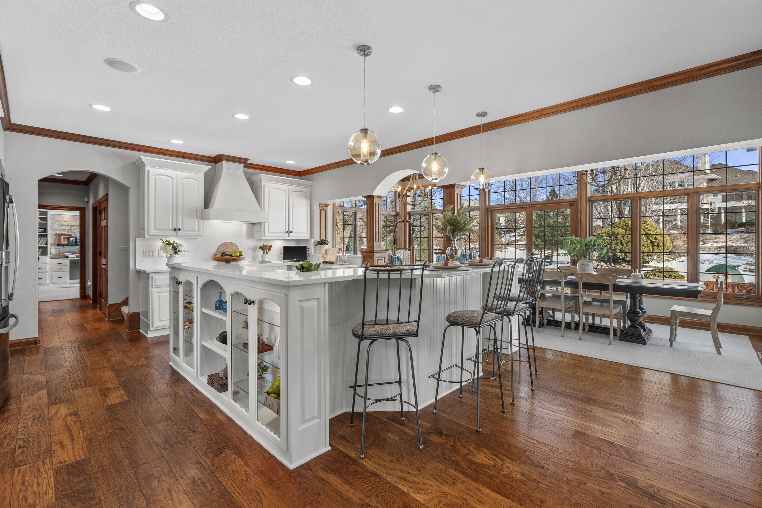 A bright kitchen and dining area with large windows, white cabinets, and wooden floors and trim, furnished with a kitchen island, bar stools, and a dining table with chairs, holding decorative plants and tableware.