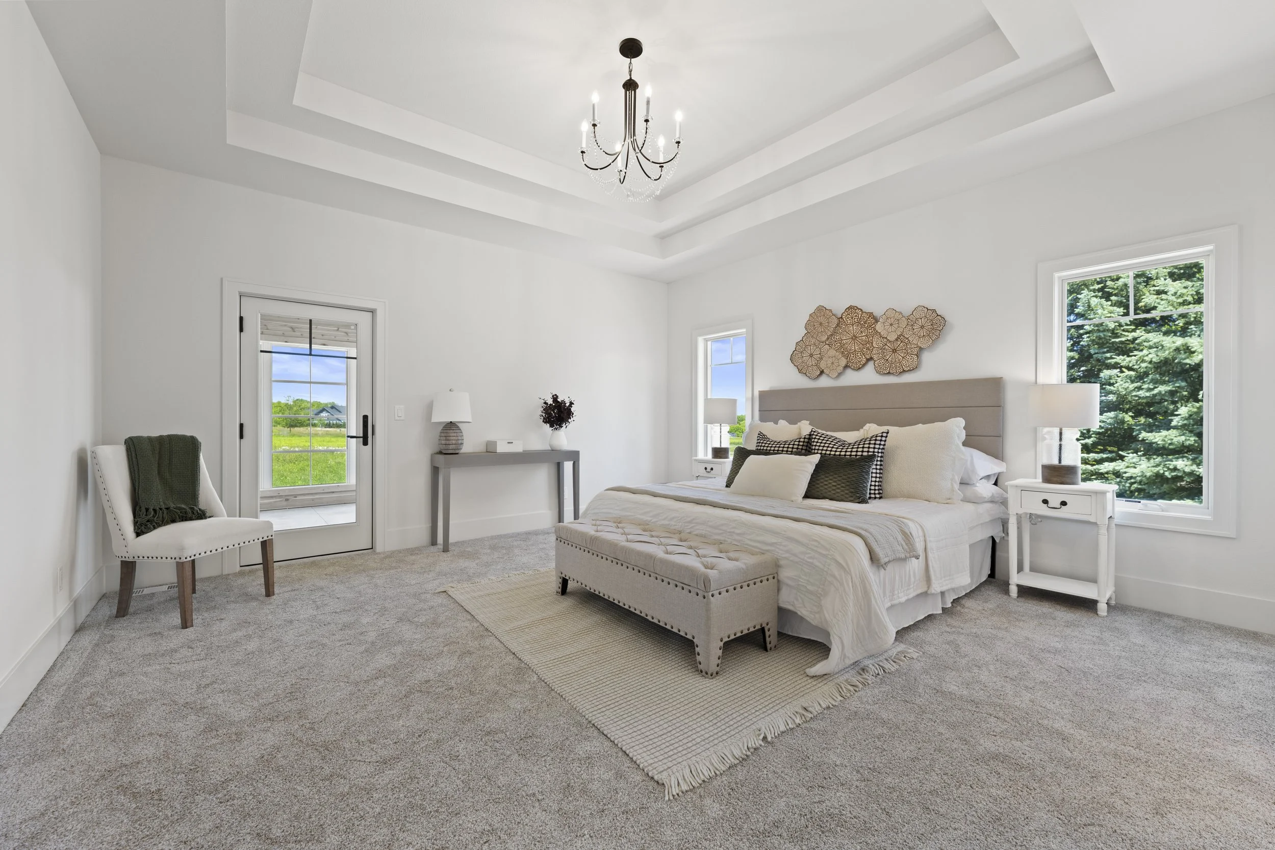 A bedroom with white walls and ceiling, featuring a chandelier and two windows showing green trees outside. The bed has beige and black pillows, a beige upholstered bench at the foot, and matching white nightstands with lamps. There is a decorative w