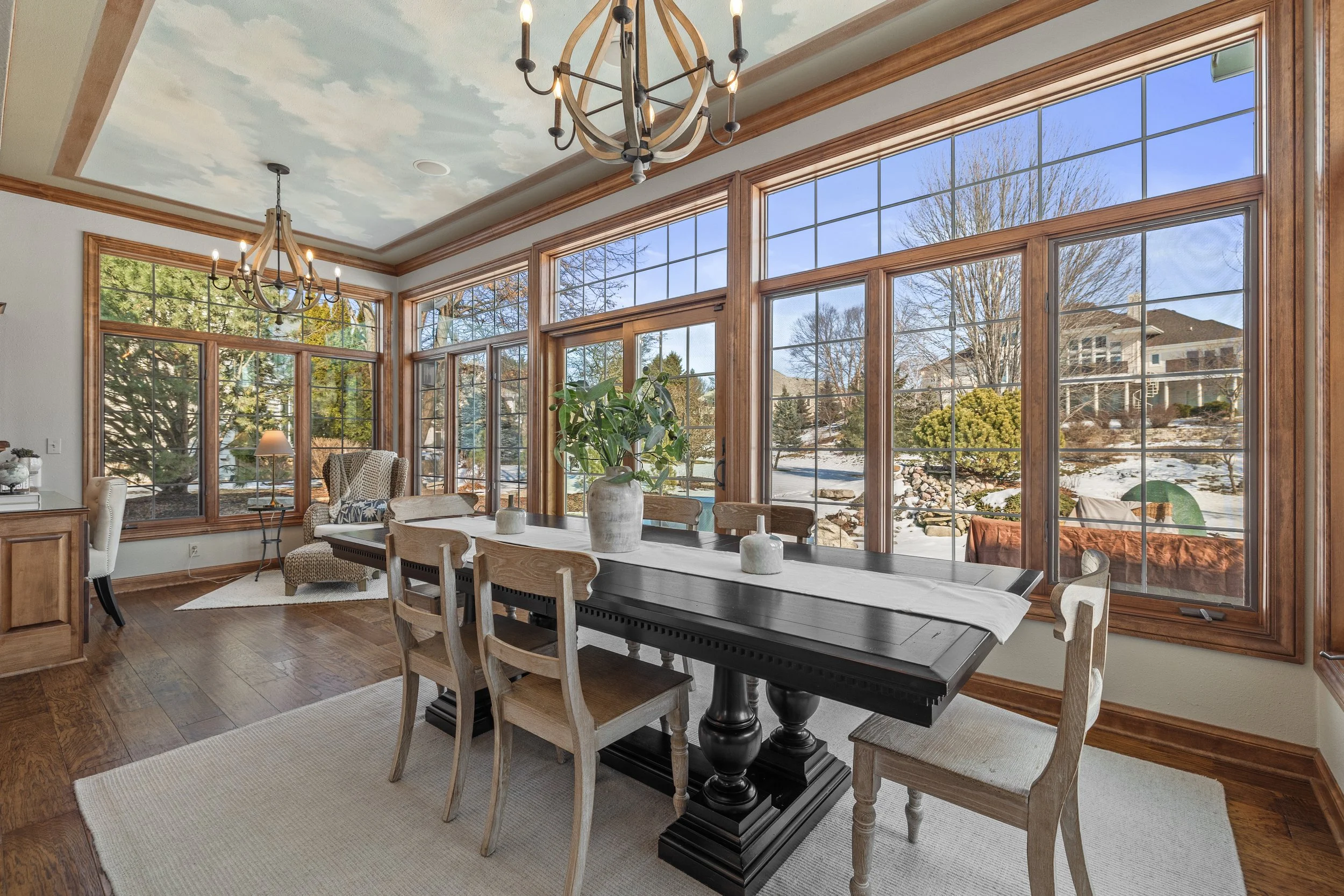 Bright dining room with large windows, wooden trim, and a ceiling painted to resemble a cloudy sky. A black dining table with a white runner is surrounded by wooden chairs with light cushions. Decorative vases and a green plant are on the table. Two 