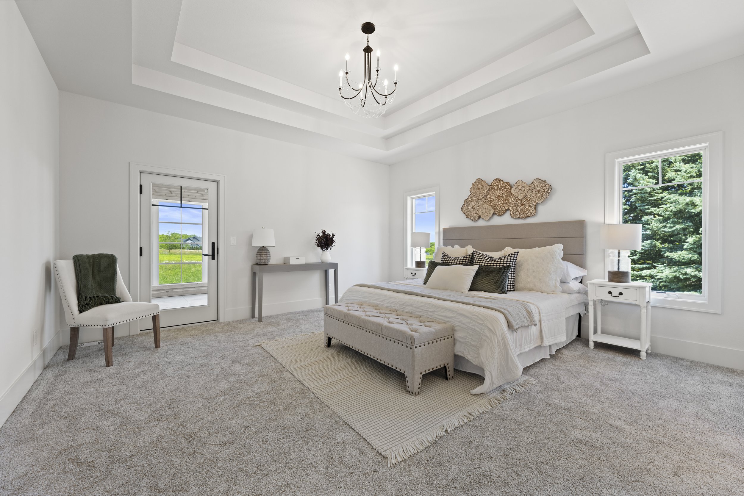 Bright bedroom with white walls, a bed with neutral linens and multiple pillows, two nightstands with lamps, a small table with a vase, a white chair with a green blanket, and large windows showing green trees and blue sky.