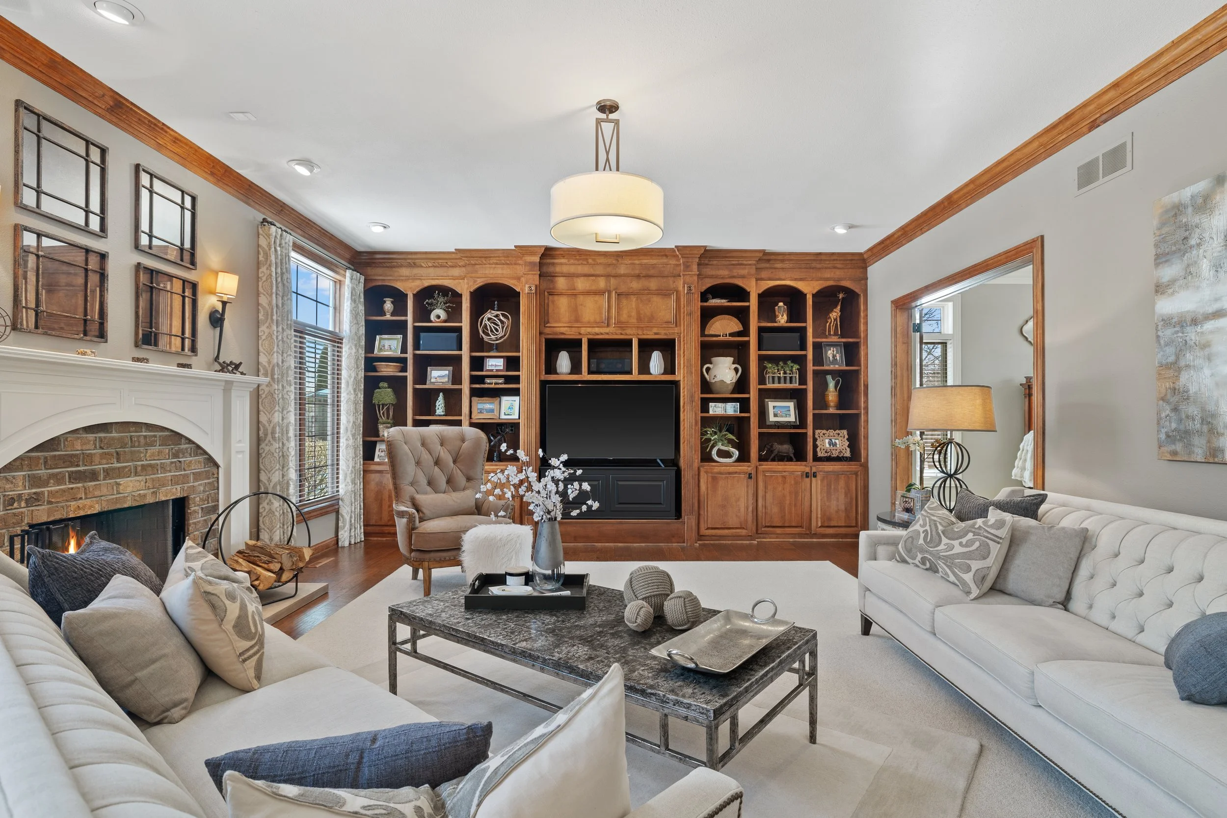Cozy living room with cream sofas, a fireplace with brick, a large wooden bookshelf, decorative objects, framed art, and a window with patterned curtains.