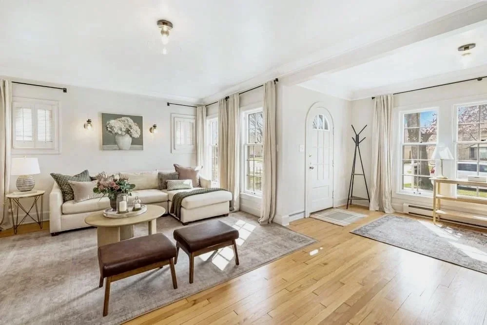 Bright living room with a cream sectional sofa, coffee table with flowers, wall art, side tables, and large windows with curtains.