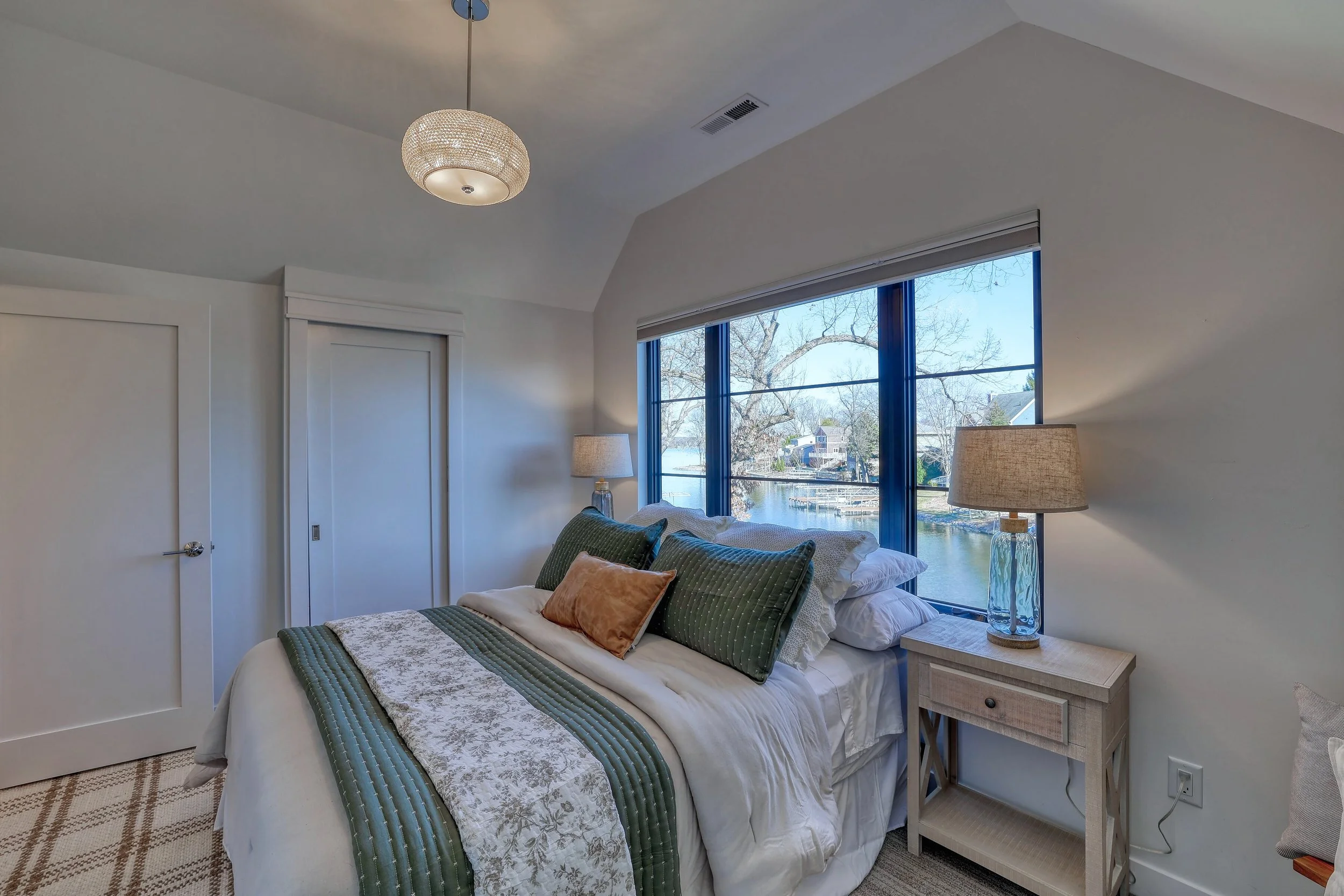 Bedroom with large window showing trees and houses outside, bed with green and white bedding, multiple pillows, and two table lamps, one on each side of the bed.
