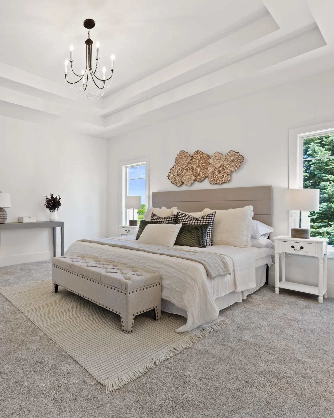 A bright bedroom with a large bed, beige upholstered headboard, and white bedding with multiple pillows. A beige tufted bench is at the foot of the bed. There are two white nightstands with lamps, and a gray console table with a vase of flowers. Two windows show greenery outside. The ceiling has layered tray details with a black chandelier hanging from the center, and the flooring is covered with a light gray carpet and a beige rug.
