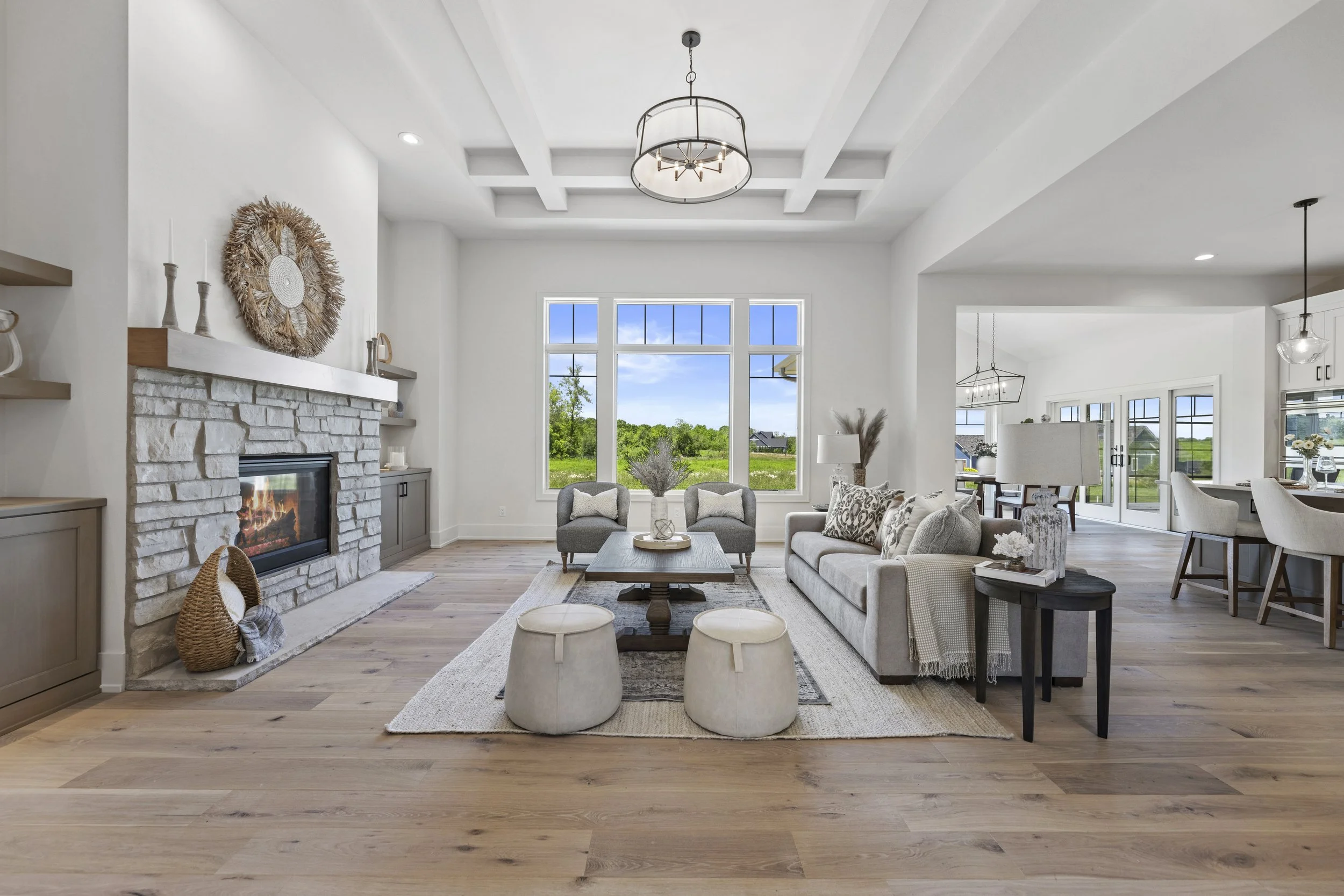 Bright living room with white walls and large windows showing a green outdoor landscape. There is a white brick fireplace with a wooden mantel decorated with candles, vases, and a large decorative wreath. The room features seating including a gray armchair, a beige sofa with patterned pillows, and three small ottomans. A wooden coffee table is in the center, topped with a decorative tray and vase. The ceiling has a coffered design with a modern chandelier. The open space flows into a dining area with a table and chairs, and French doors lead to an outdoor deck.
