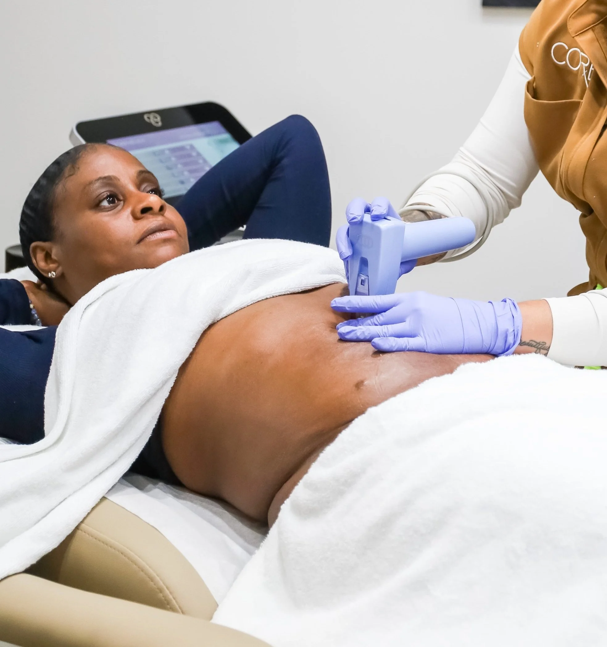 A woman receiving a non-invasive body treatment from a professional using a handheld device, with another person providing care. The person receiving the treatment is lying down with a towel over her legs.