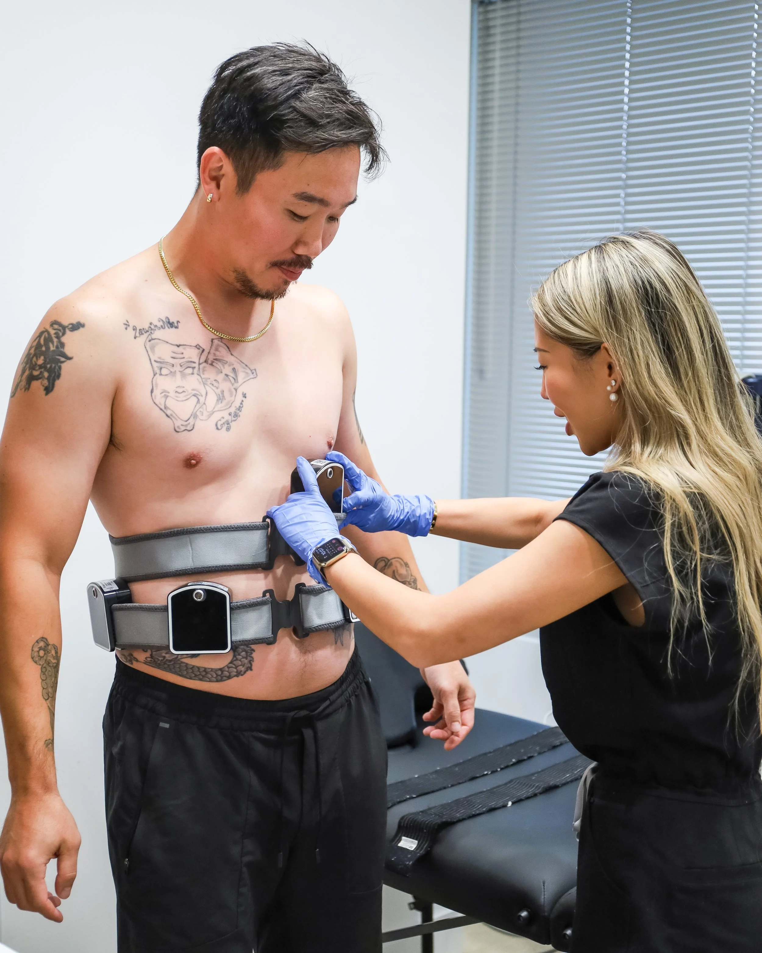 A man standing shirtless while a woman wearing gloves adjusts a medical device around his torso.