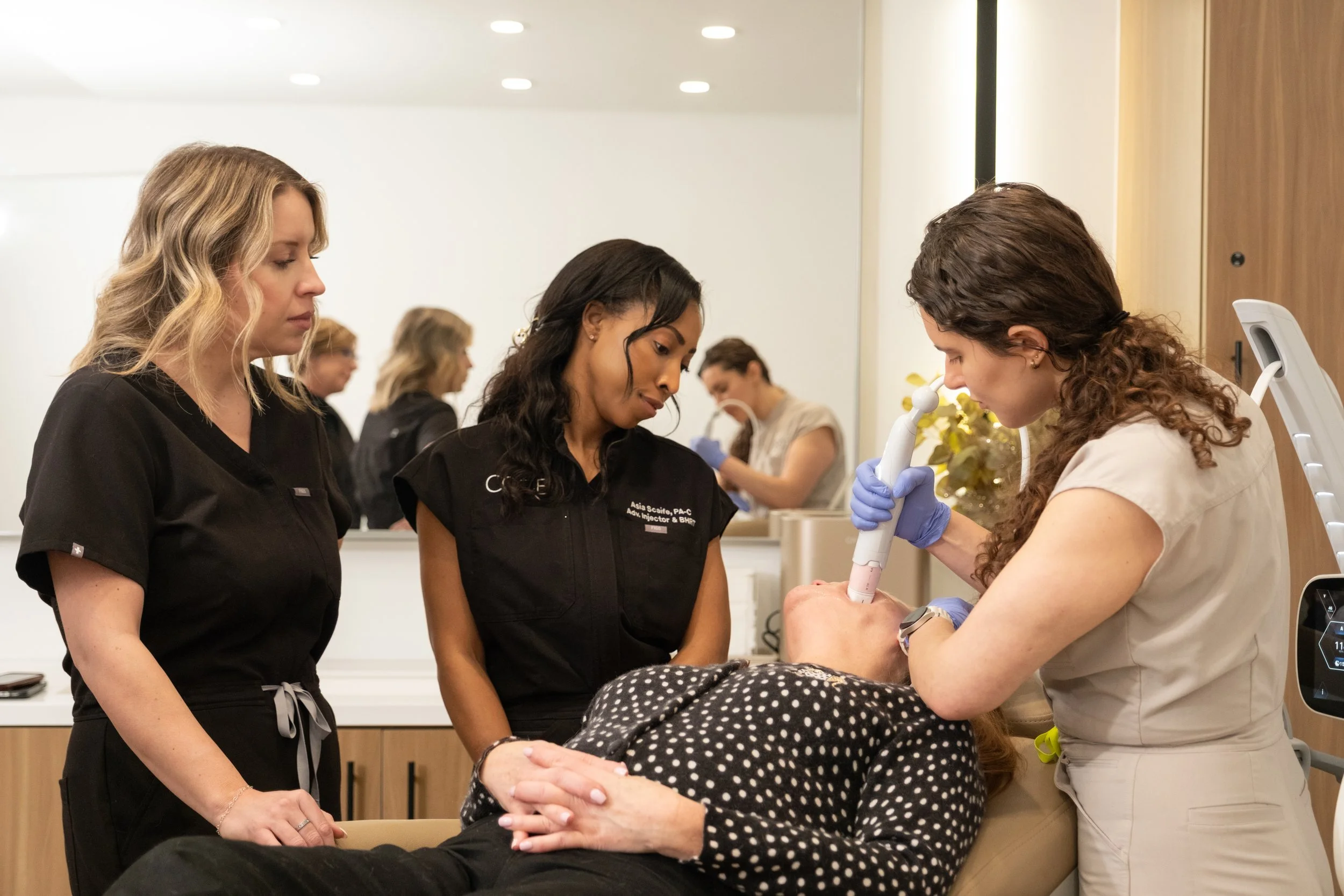 A woman sitting in a chair looking at herself in a hand mirror, with another person standing nearby, in a medical or cosmetic consultation setting.