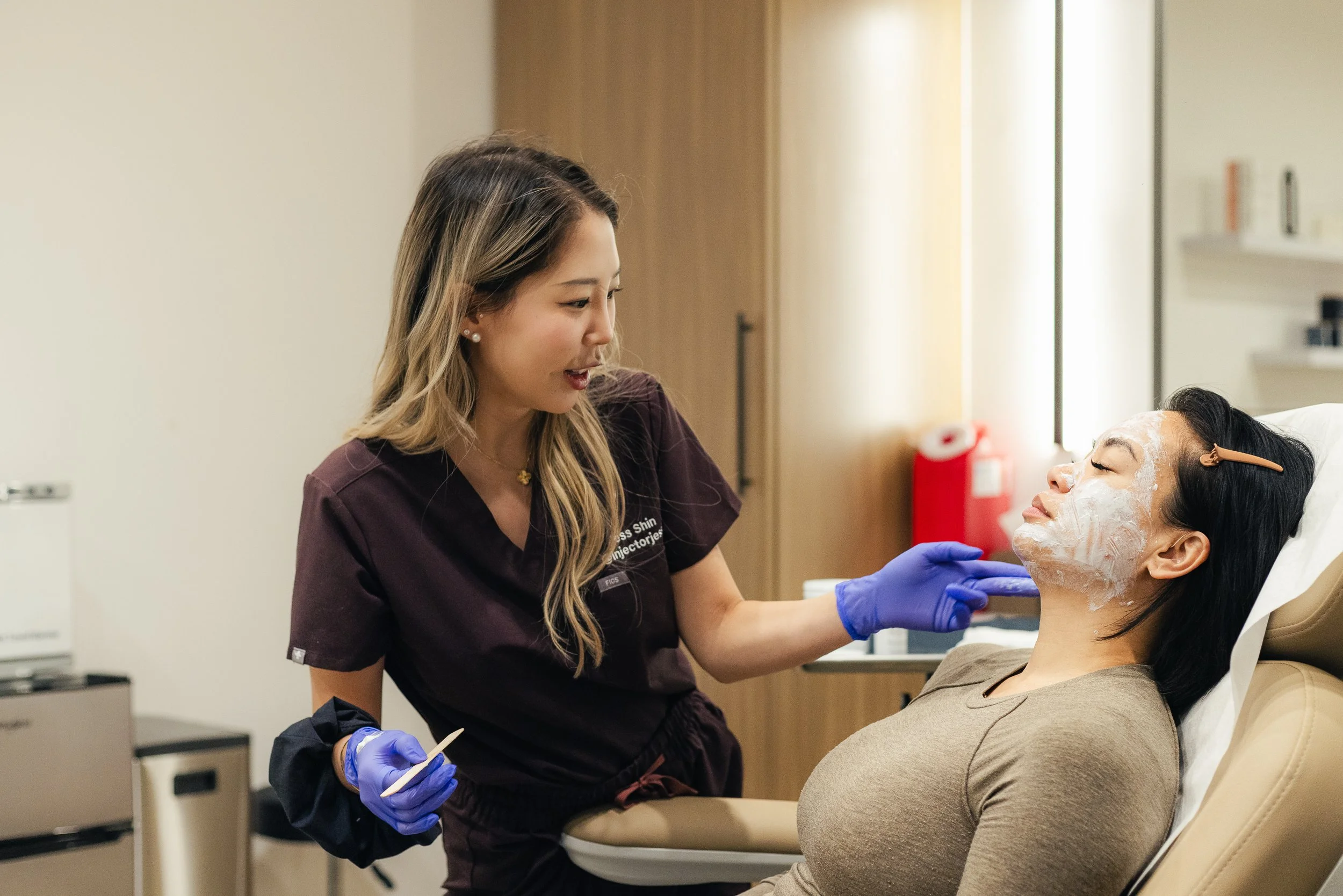 A woman sitting in a chair looking at herself in a hand mirror, with another person standing nearby, in a medical or cosmetic consultation setting.