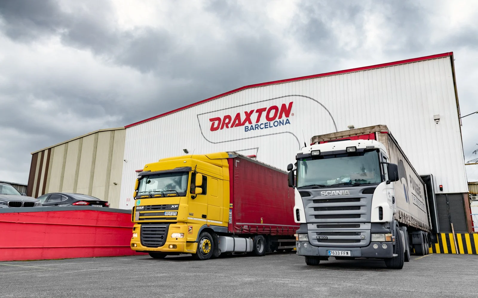 Nave con rótulo “DRAXTON BARCELONA” y dos tráileres cargando en el muelle de carga.