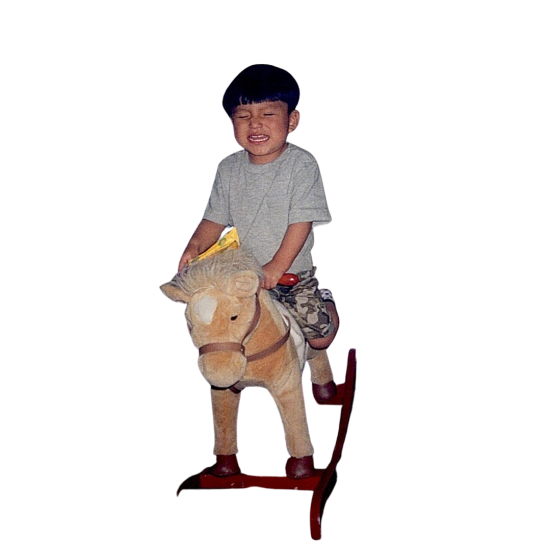 Child riding a rocking horse with eyes closed, wearing a gray shirt and camo shorts, against a plain background.