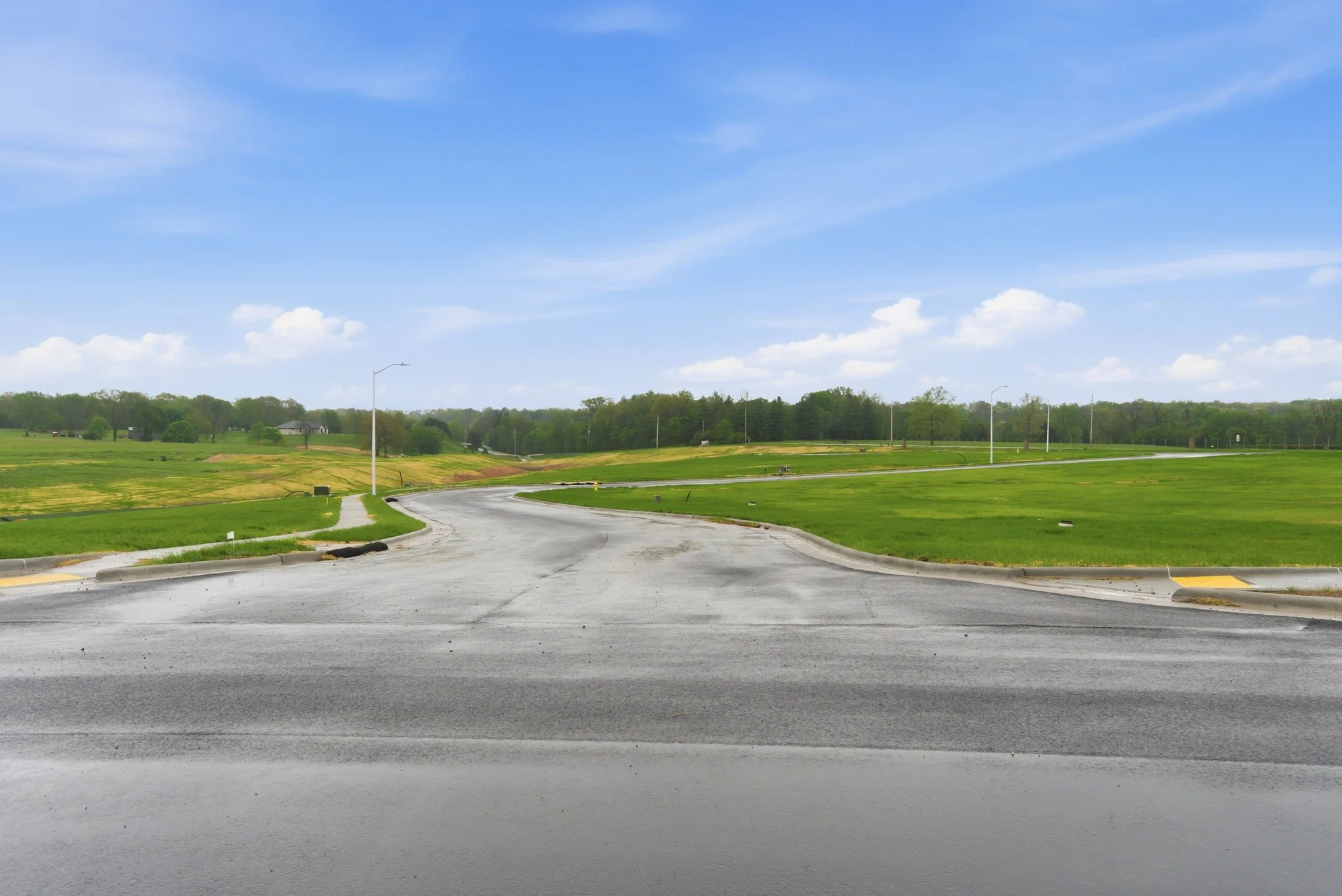 An empty curved road in a rural area with green grass and trees in the background under a partly cloudy blue sky.