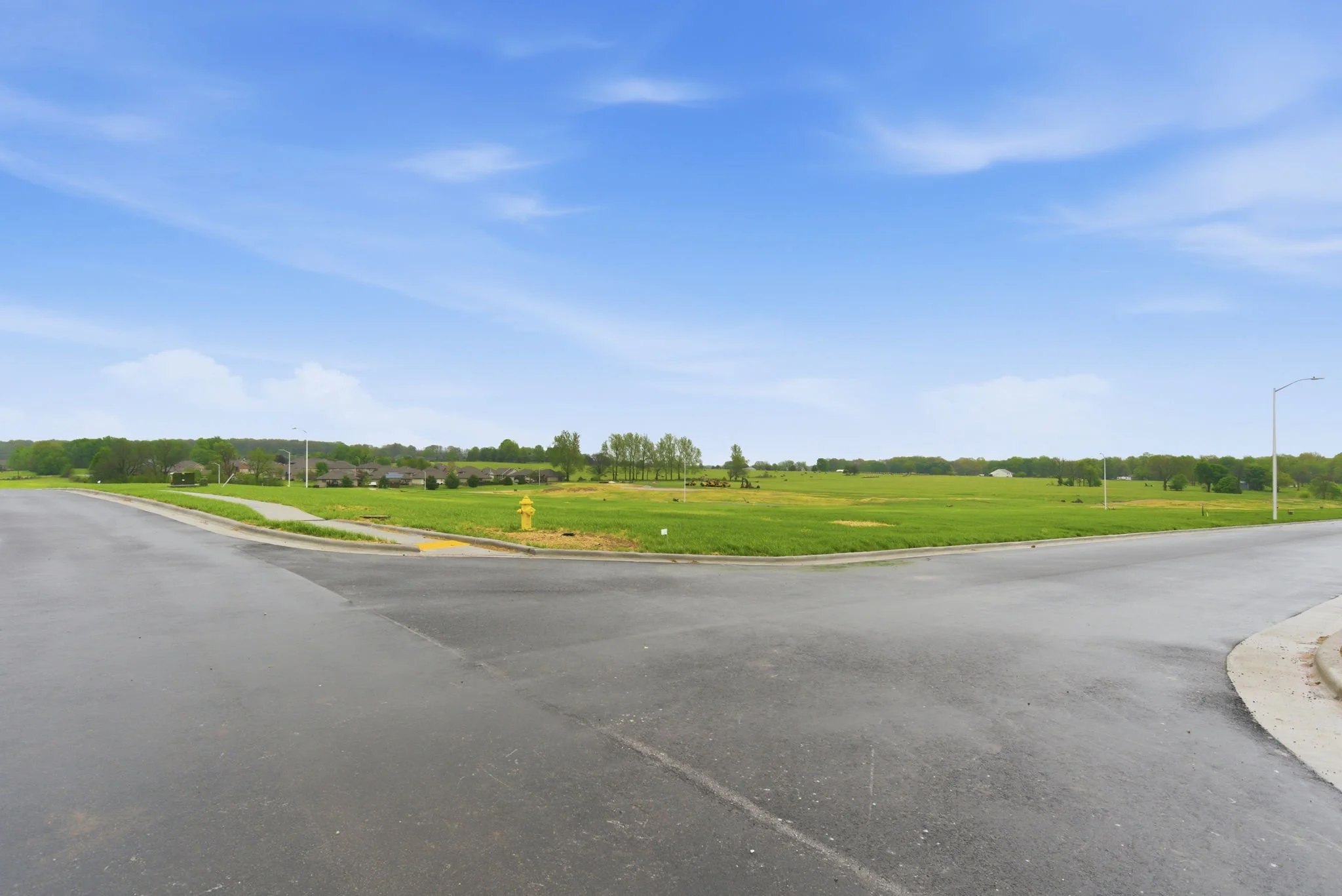 A suburban scene with a paved street corner, a grassy field, houses in the distance, and a clear blue sky with scattered clouds.